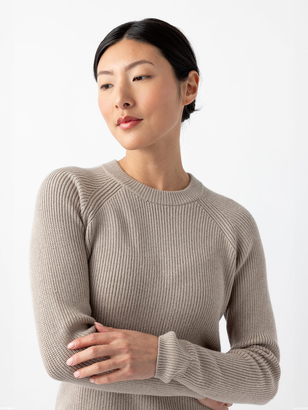 A person with dark hair pulled back is wearing a light gray, long-sleeved Women's Classic Crewneck from Cozy Earth. They are looking to the side with a neutral expression and have their arms crossed gently in front of them. The background is plain white. |Color:Taupe