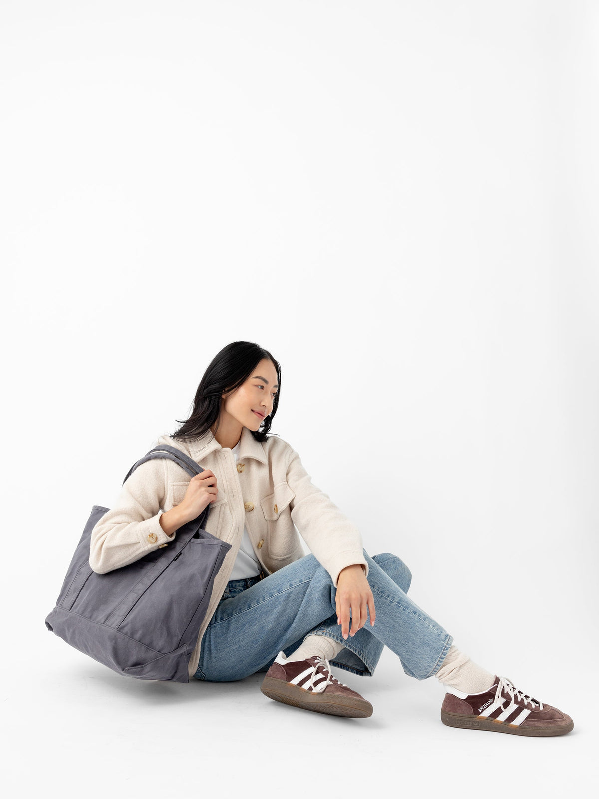 A woman dressed in a cream-colored sweater and blue jeans sits on the ground with her legs crossed. She holds a large Waxed Canvas Tote from Cozy Earth and is wearing brown sneakers. The background is a plain white studio setting. |Color:Washed Navy