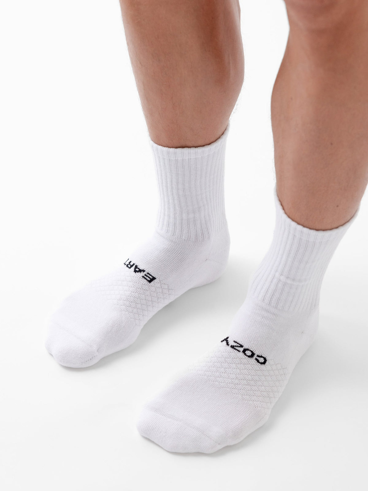 A person wears white Essential Calf Socks from Cozy Earth; the left sock reads "EARTH" and the right "COZY" in black letters. The background is plain white. |Color:White