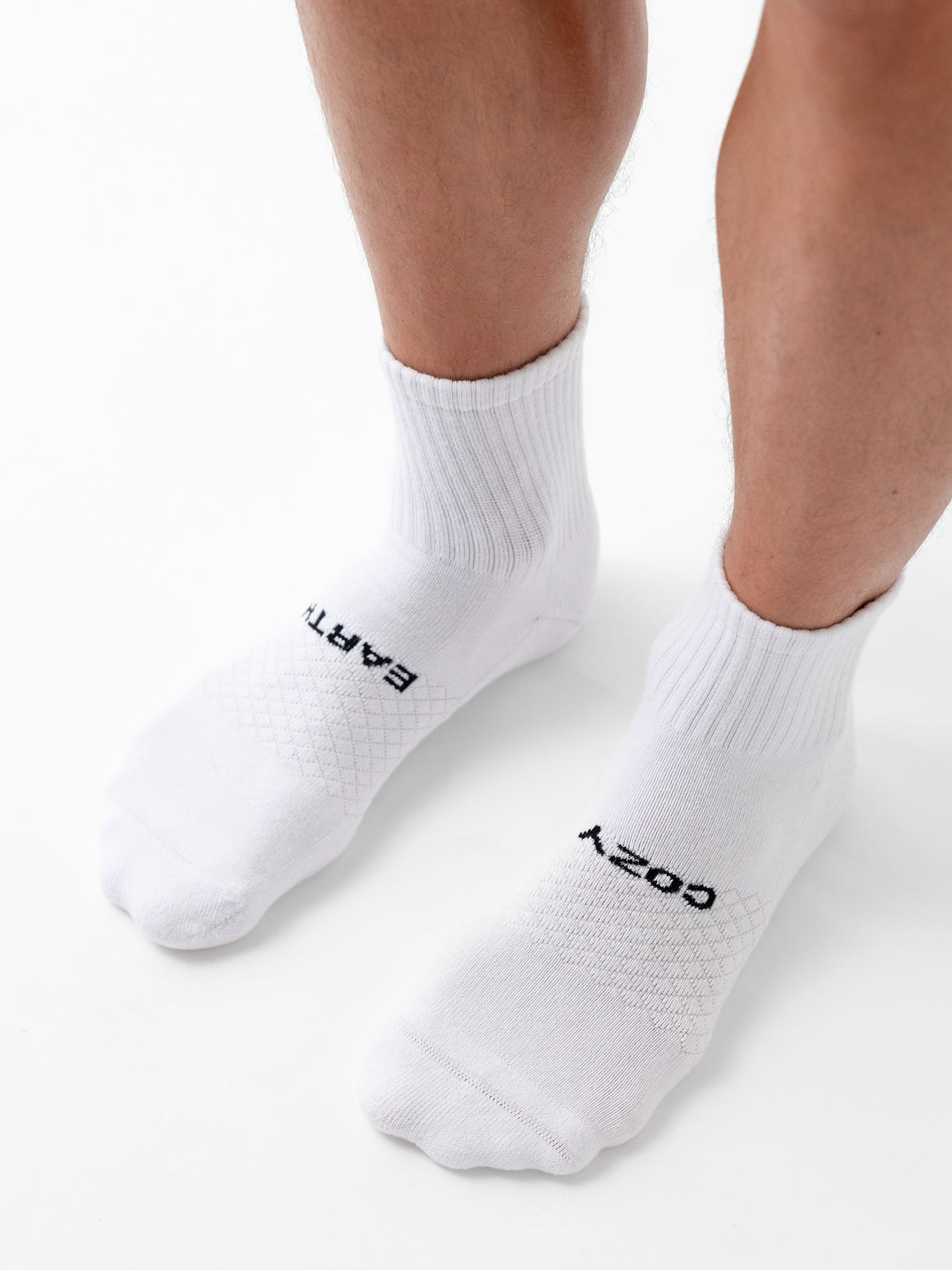 Someone stands on a white surface wearing Cozy Earth’s Essential Quarter Socks featuring ribbed cuffs and black "EARTH" text on the left sock and "COZY" on the right. |Color:White