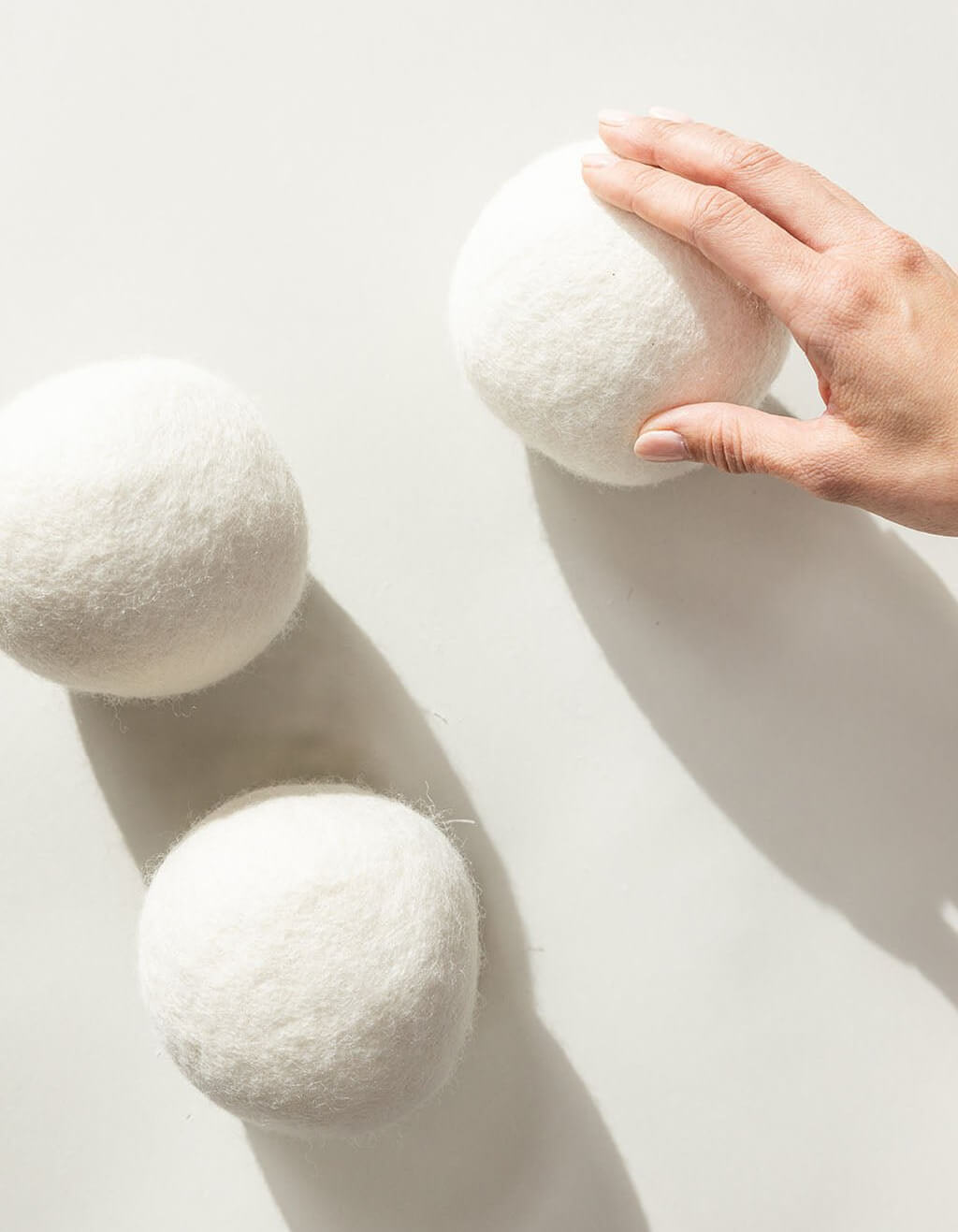 A hand touches one of three large, white Cozy Earth Wool Dryer Balls resting on a light surface, with soft shadows cast beneath them.|Size:Large