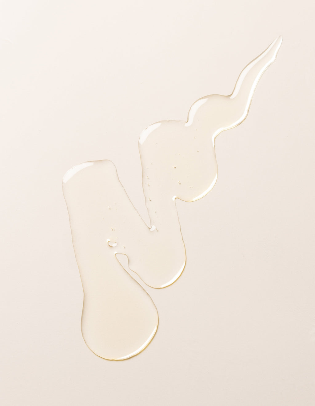 A swirl of transparent, light-colored Cozy Earth's Revitalizing Pair liquid on a plain, pale beige surface. The liquid forms irregular shapes with droplets at the ends, creating a visually interesting pattern.