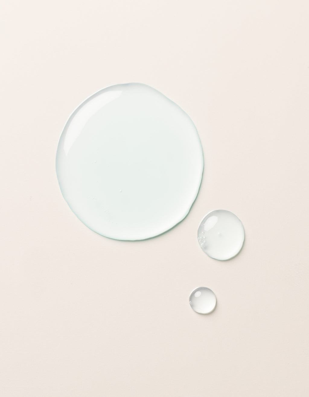 A large clear droplet and two smaller droplets of Cozy Earth Hydrating Serum rest on a smooth, light beige surface. The glossy, semi-transparent droplets reflect light slightly on their surfaces.