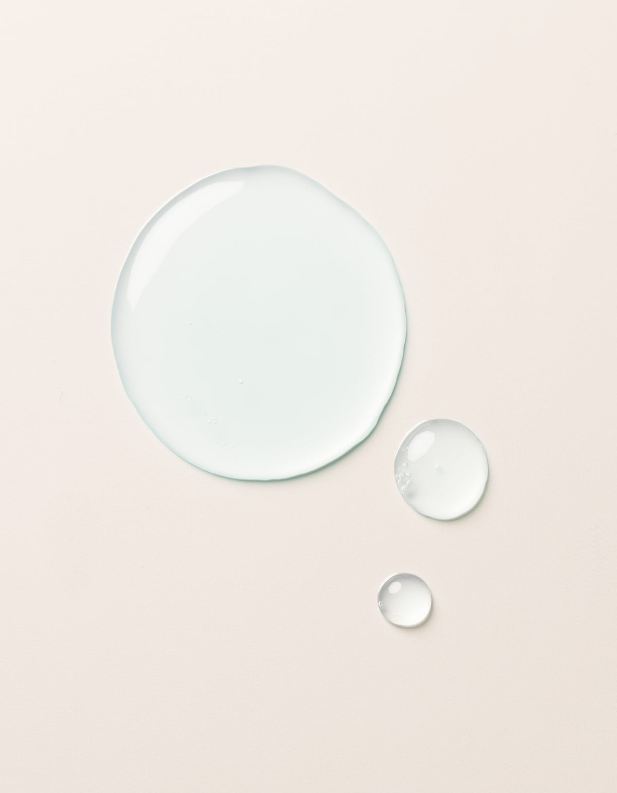 A large clear droplet and two smaller droplets of Cozy Earth Hydrating Serum rest on a smooth, light beige surface. The glossy, semi-transparent droplets reflect light slightly on their surfaces.