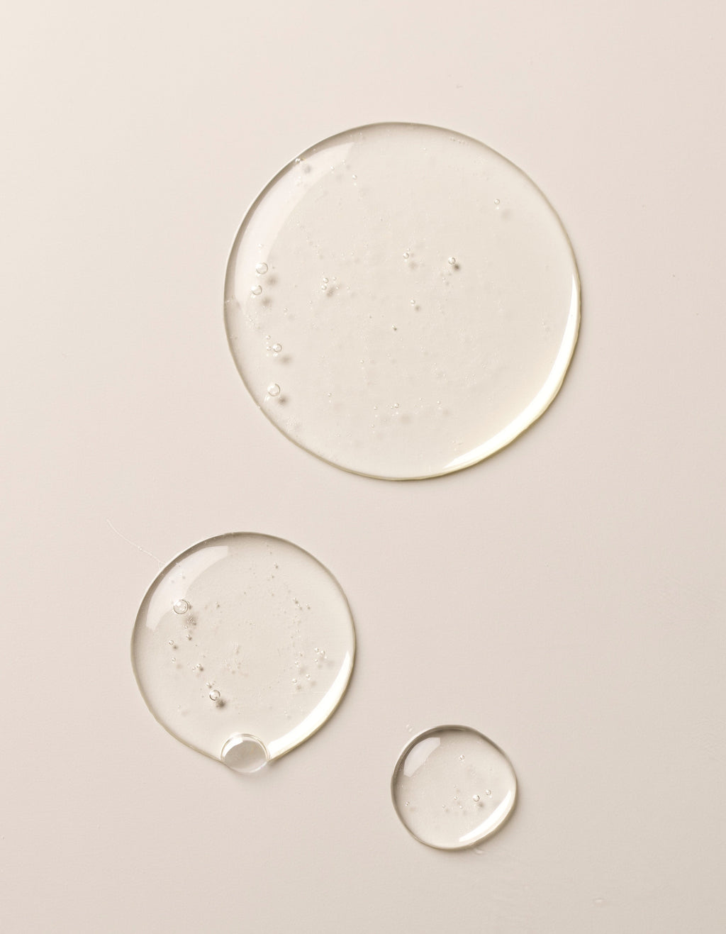 Three clear drops of Gel Cleanser by Cozy Earth on a beige surface. The drops vary in size, ranging from a large drop at the top to smaller ones below, showcasing a glossy and slightly reflective texture.|Size:Full Size