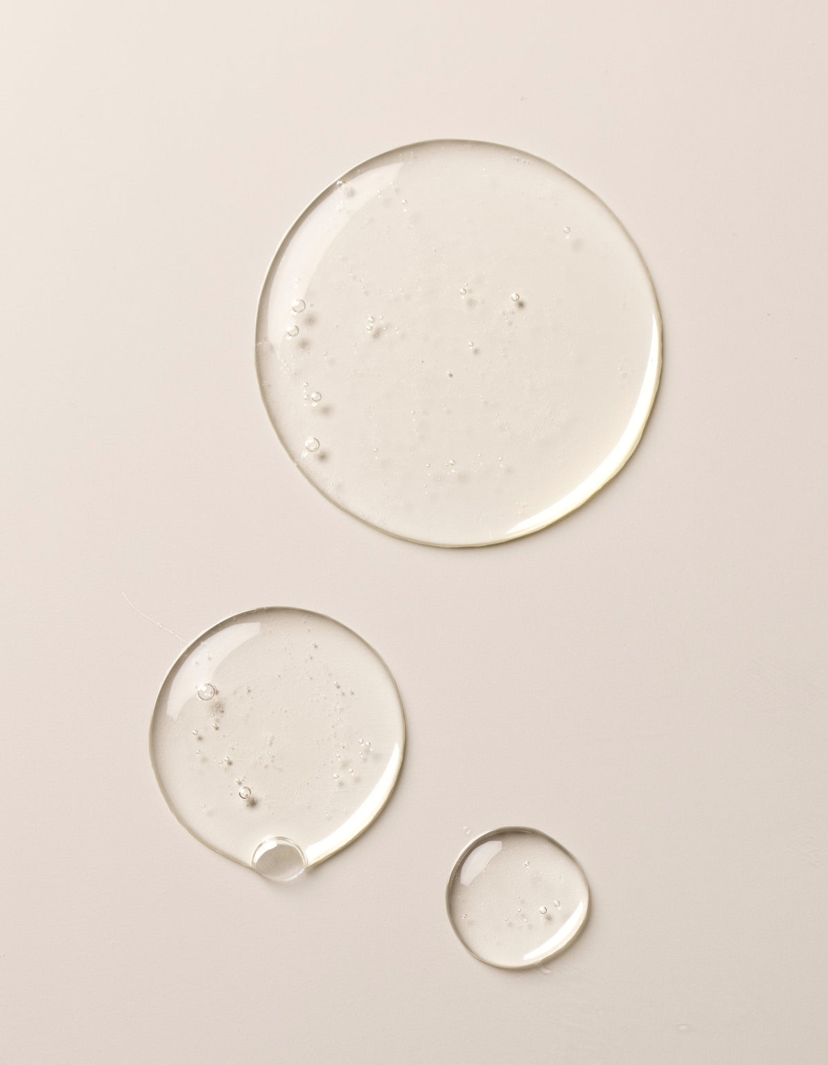 Three clear drops of Gel Cleanser by Cozy Earth on a beige surface. The drops vary in size, ranging from a large drop at the top to smaller ones below, showcasing a glossy and slightly reflective texture.|Size:Full Size