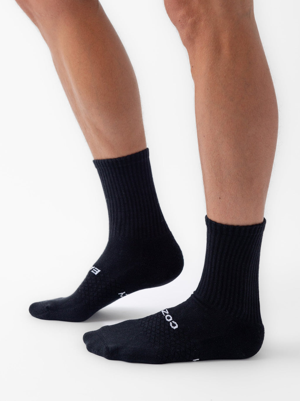 A person wearing Cozy Earth Essential Calf Socks stands on a white background, showing both legs from the knee down. The black socks have a ribbed texture and white text on the top of the foot. |Color:Black