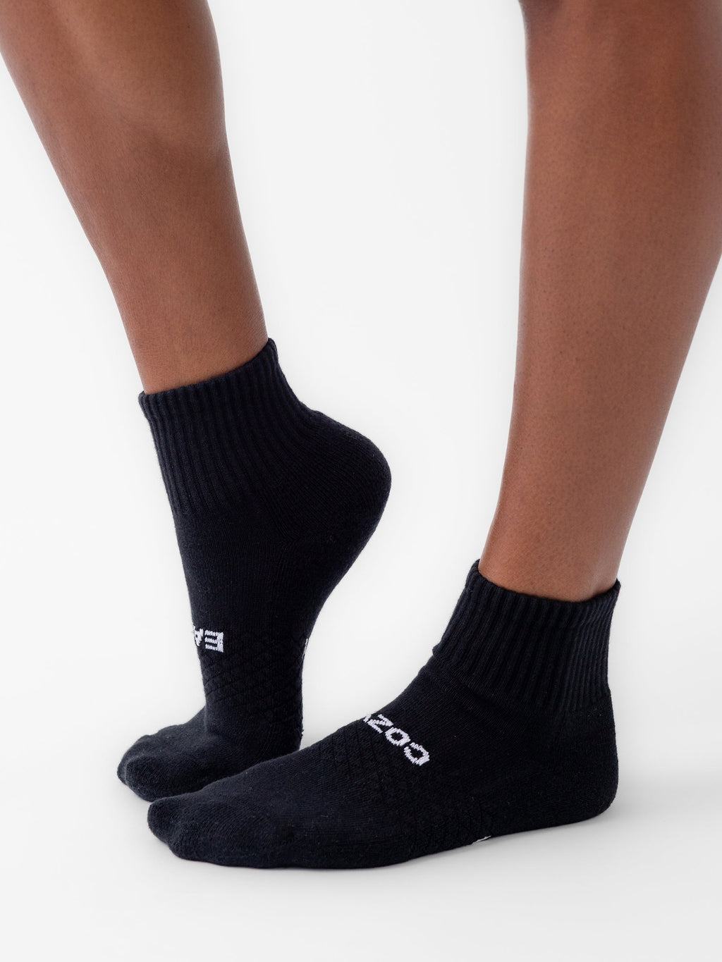 A person wears Cozy Earth's Essential Quarter Sock (6-Pack) in black, featuring a ribbed cuff, textured pattern, and white text near the toes. Only the lower legs and feet are visible as they stand on a white surface. |Color:Black