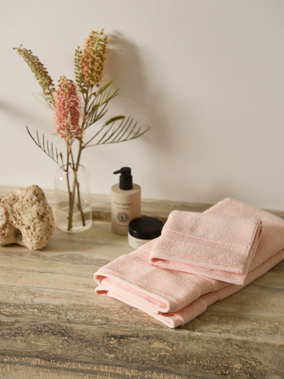 Cozy Earth's Luxe Bath Towel Set is neatly folded on a marble countertop next to a lotion bottle and a small jar. A clear vase with dried flowers enhances the decor, while a nearby textured stone adds to the natural aesthetic. |Color:Blossom