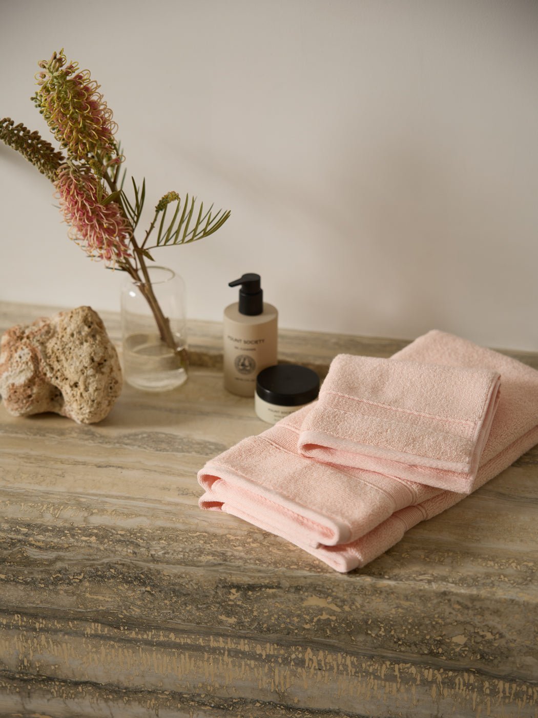 The Complete Luxe Bath Bundle by Cozy Earth features soft pink towels on a marble countertop, complemented by a lotion bottle, small jar, decorative coral, and vase with pink flowers and greenery. |Color:Blossom