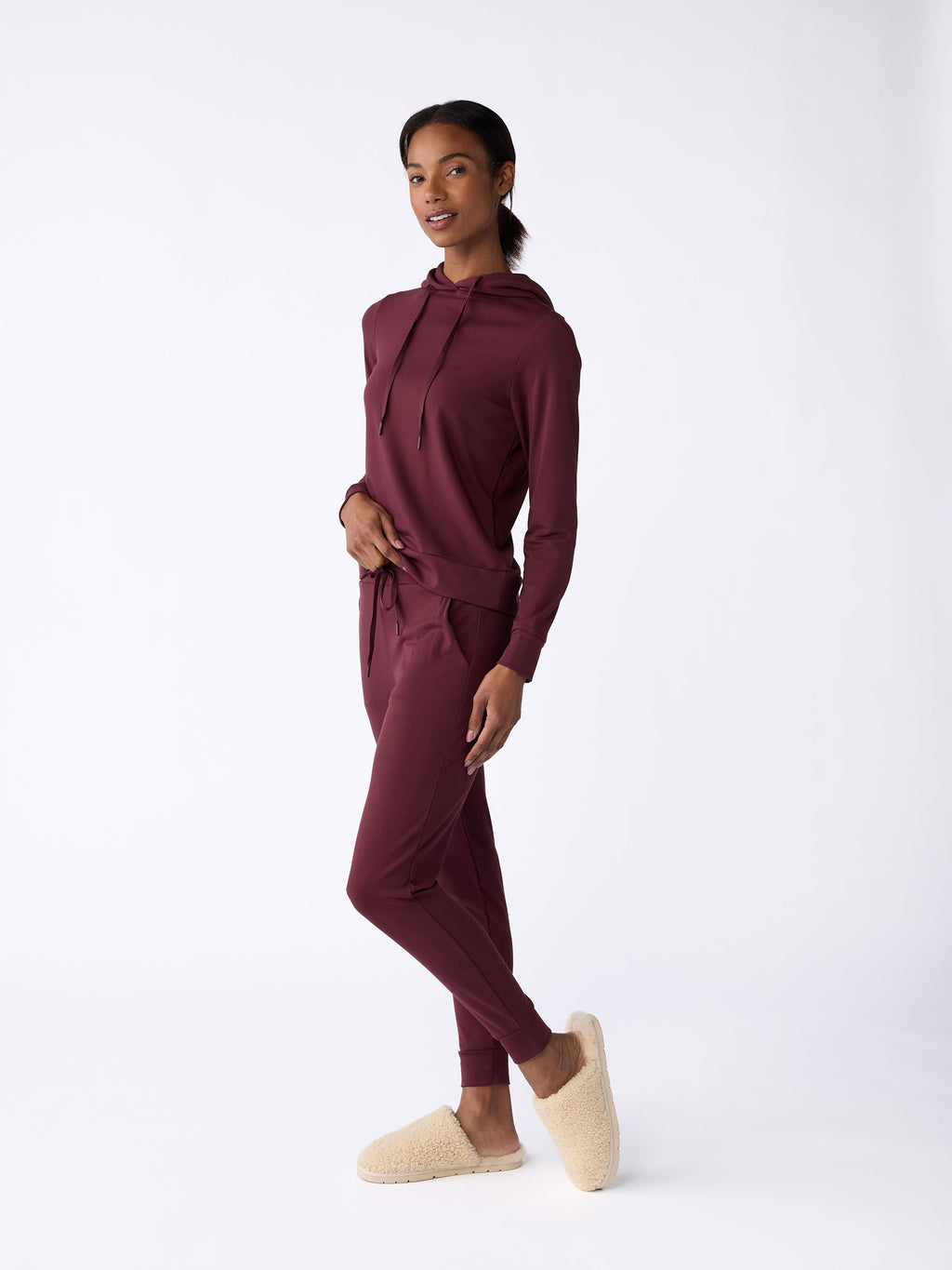 A person smiling, dressed in a maroon hoodie and Cozy Earth's Women's Bamboo Jogger Pant, poses confidently against a white background. They complete the look with cozy, beige slippers. |Color:Burgundy