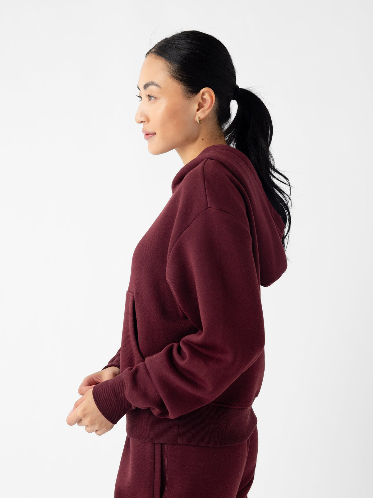 A person with long dark hair tied in a ponytail is seen in a profile view, wearing the Cozy Earth Women's CityScape Cropped Hoodie in maroon and matching pants. The background is plain white, and the person is looking to the left with a relaxed expression. |Color:Burgundy