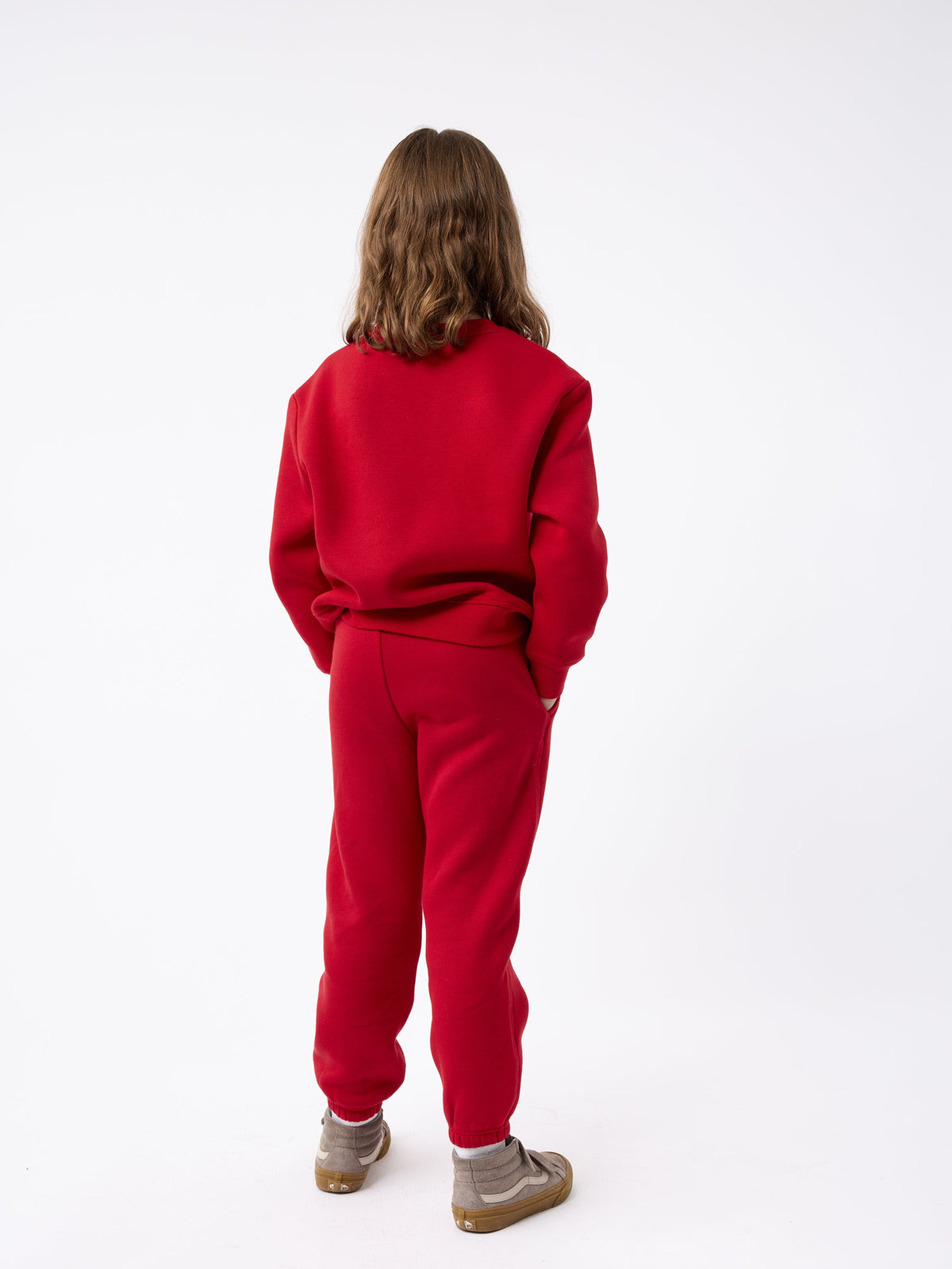 A child with medium-length brown hair is standing with their back to the camera, wearing a Kid's CityScape Crewneck by Cozy Earth in red and matching sweatpants, along with brown shoes. The background is plain white. |Color:Crimson