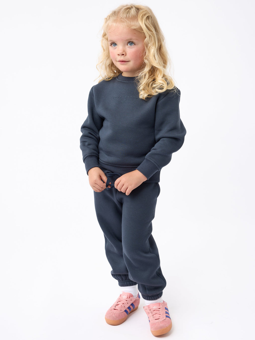 A young child with long blonde hair stands against a white background, wearing a Cozy Earth Kid's CityScape Crewneck and matching dark blue sweatpants. They sport pink sneakers accented with blue and white stripes and look thoughtfully to the side. |Color:Eclipse