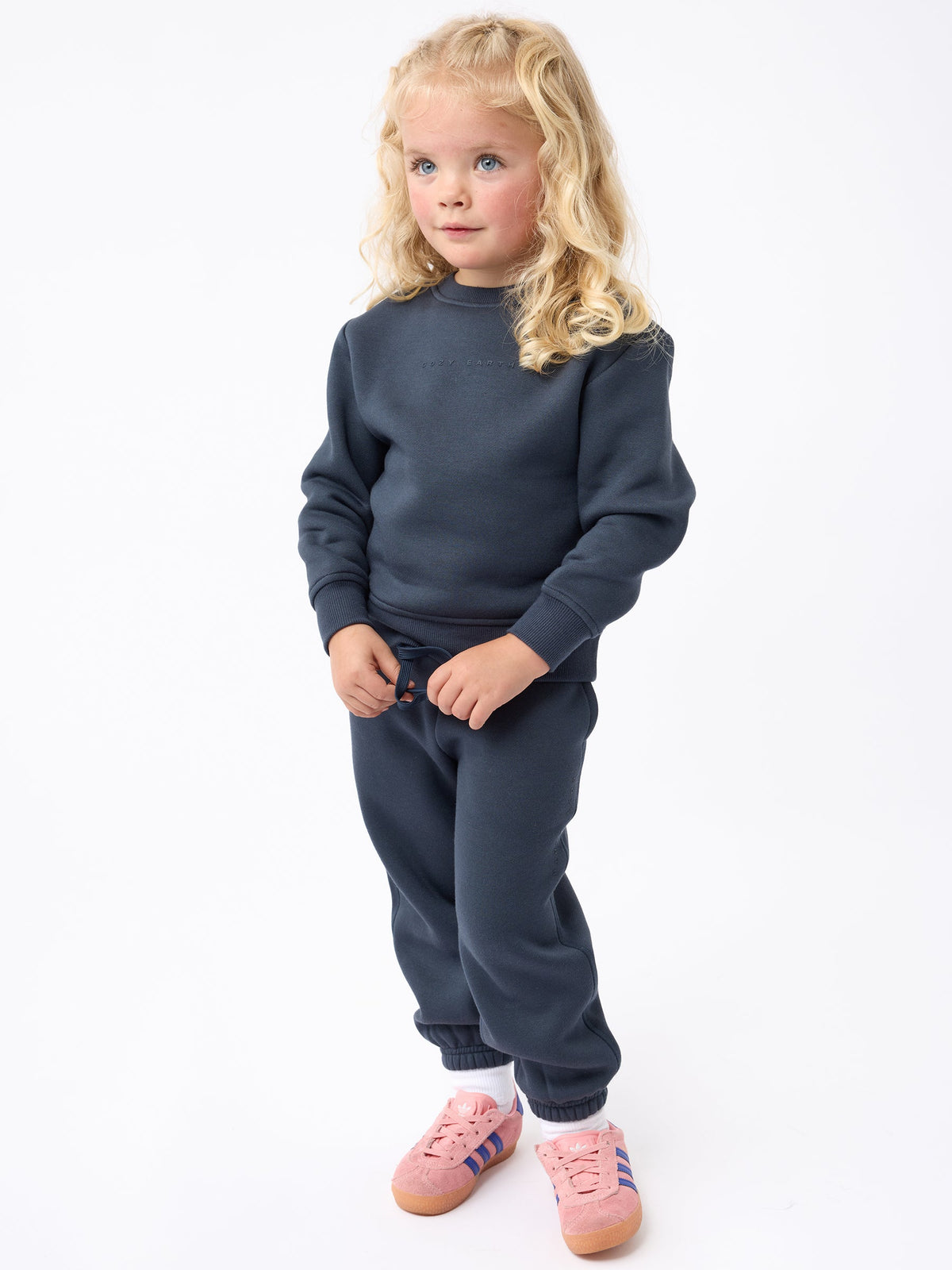 A young child with long blonde hair stands against a white background, wearing a Cozy Earth Kid's CityScape Crewneck and matching dark blue sweatpants. They sport pink sneakers accented with blue and white stripes and look thoughtfully to the side. |Color:Eclipse