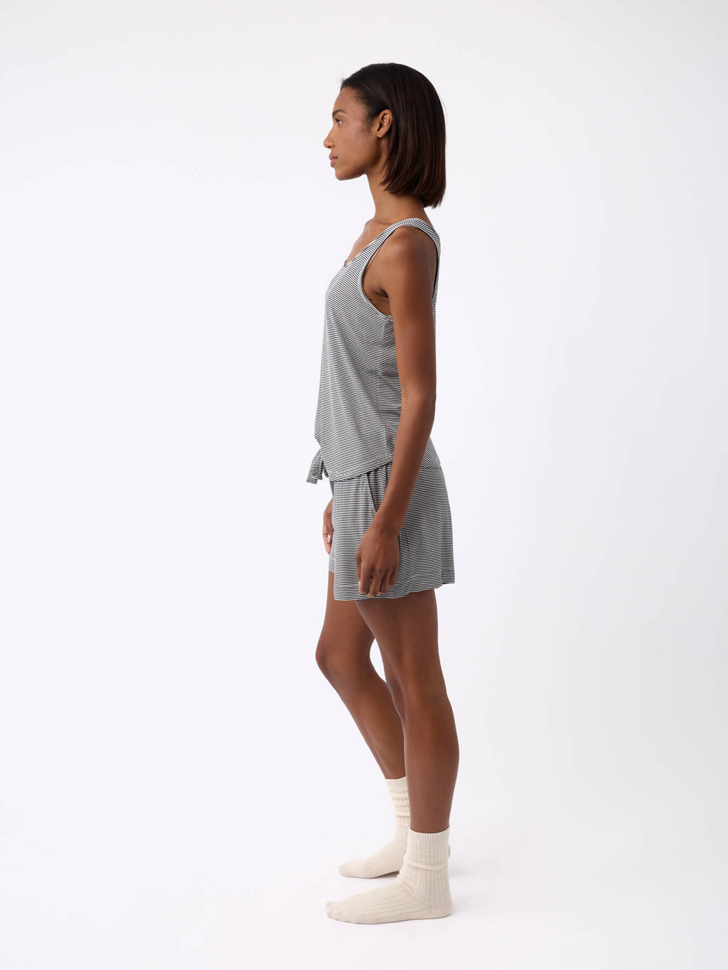 A person stands in profile against a plain white background, wearing the Women's Bamboo Stretch-Knit Lounge Tank by Cozy Earth, paired with matching shorts and cream-colored socks. |Color:Eclipse Mini Stripe