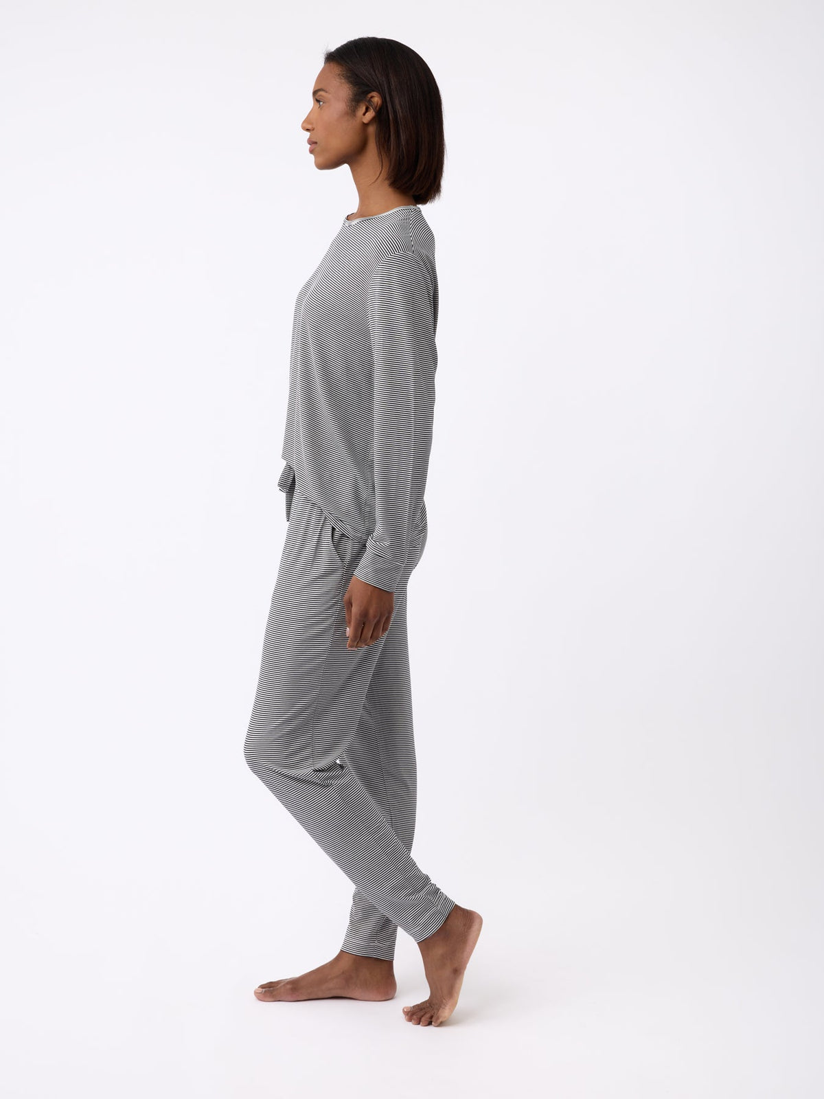 A person stands barefoot in profile, dressed in Cozy Earth's grey long-sleeve top and Women's Bamboo Stretch-Knit Tapered Pant, set against a simple white backdrop. |Color:Eclipse Mini Stripe