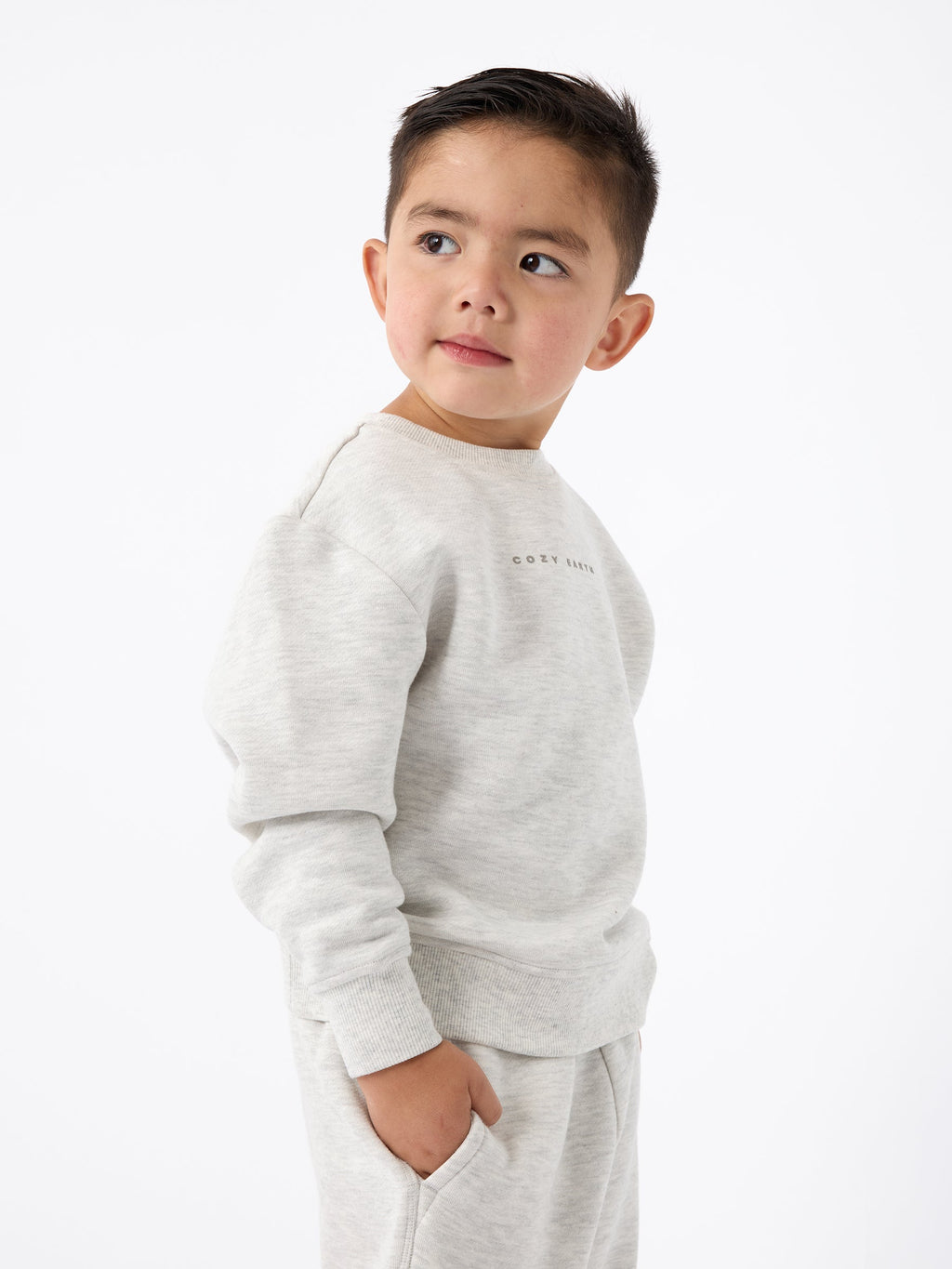 A young child with short hair is wearing Cozy Earth's light gray Kid's CityScape Crewneck sweatshirt and matching pants. They are looking to the side against a plain white background. |Color:Heather Grey