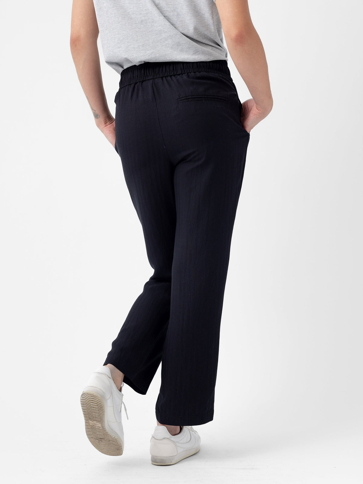 A person is shown from the back, standing against a white background. They are wearing a grey shirt and Cozy Earth's Women's Sunset Cropped Pant, which features an elastic waistband, along with white sneakers. The person's right hand is in their pocket, and they are slightly leaning back. |Color:Jet Black