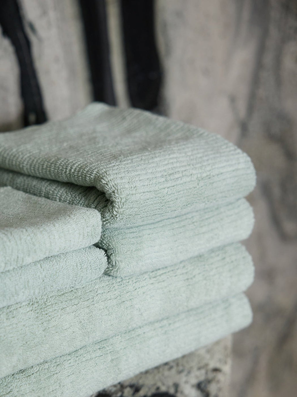 A close-up of Cozy Earth Ribbed Terry Hand Towels in light green, neatly folded on a marble surface with a softly blurred background.|Color:Laurel