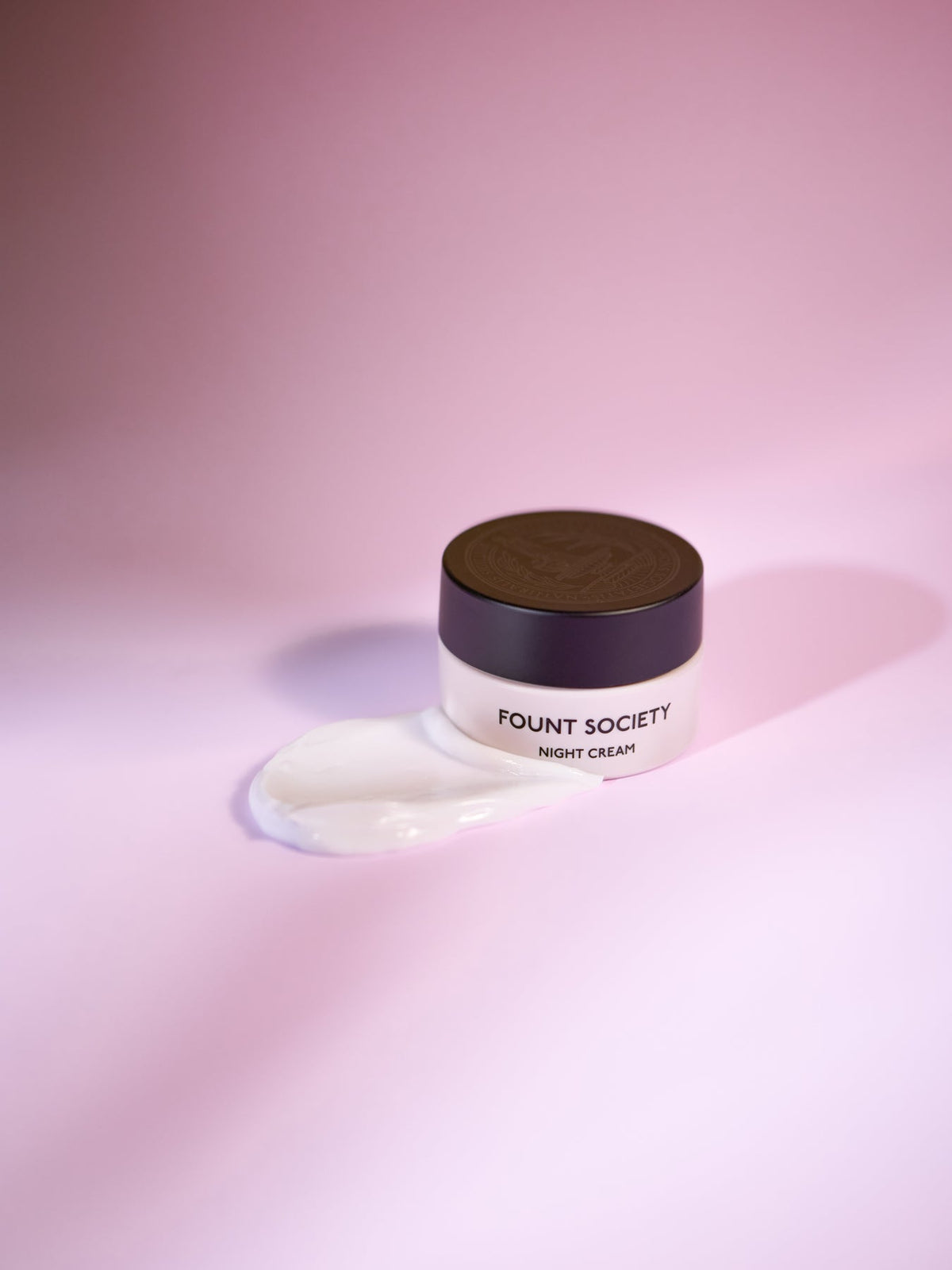 A jar of Cozy Earth Night Cream sits on a pink surface, with some of the cream spilled beside it. The jar features a dark lid that stands out against the soft background. |Size:Mini