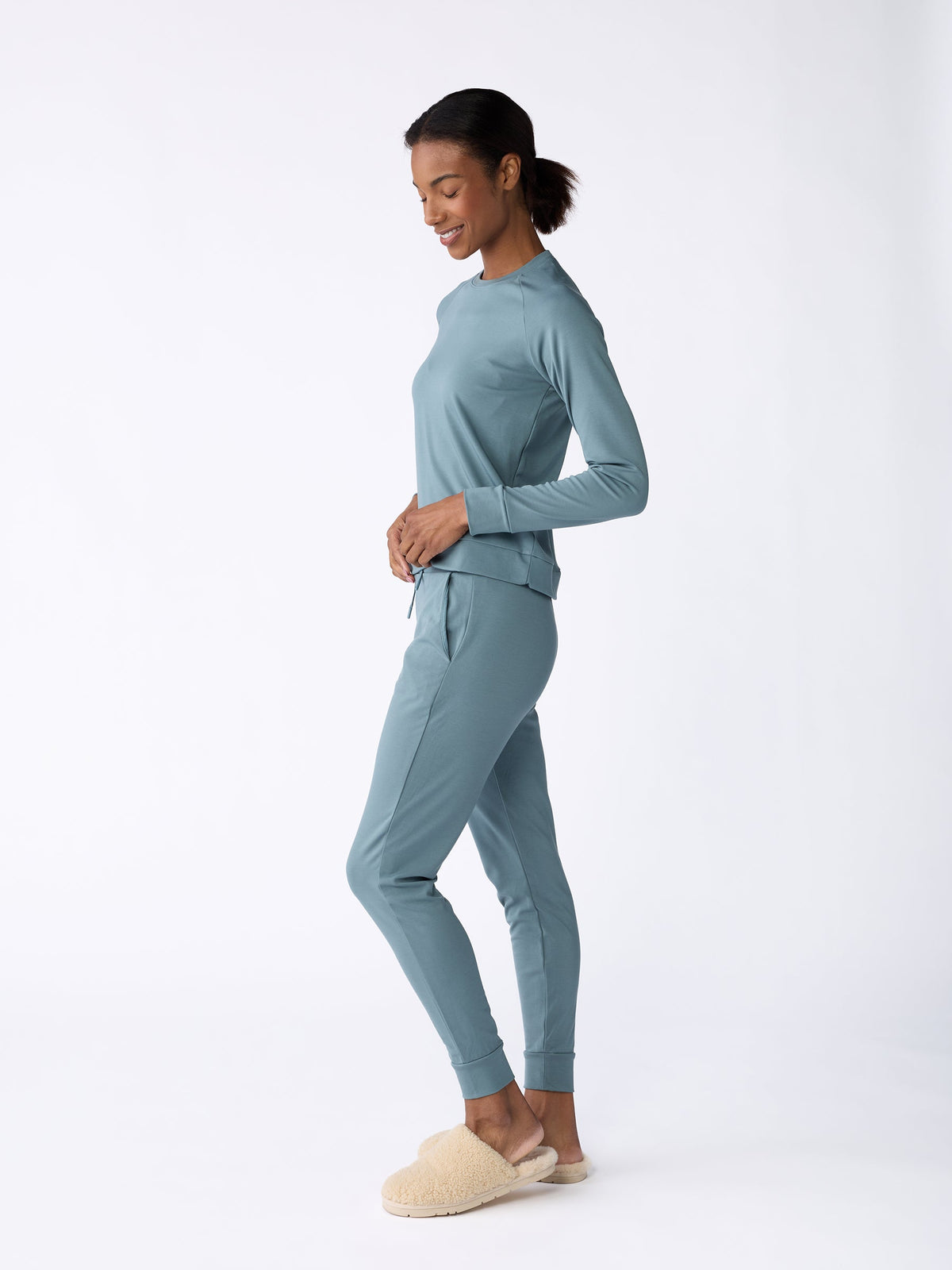 A woman wearing Cozy Earth's Women's Bamboo Jogger Pant in teal and beige slippers smiles while standing in a relaxed pose against a white background. Her hair is pulled back as she stands in profile, with her hands resting on her hips. |Color:Oceanside