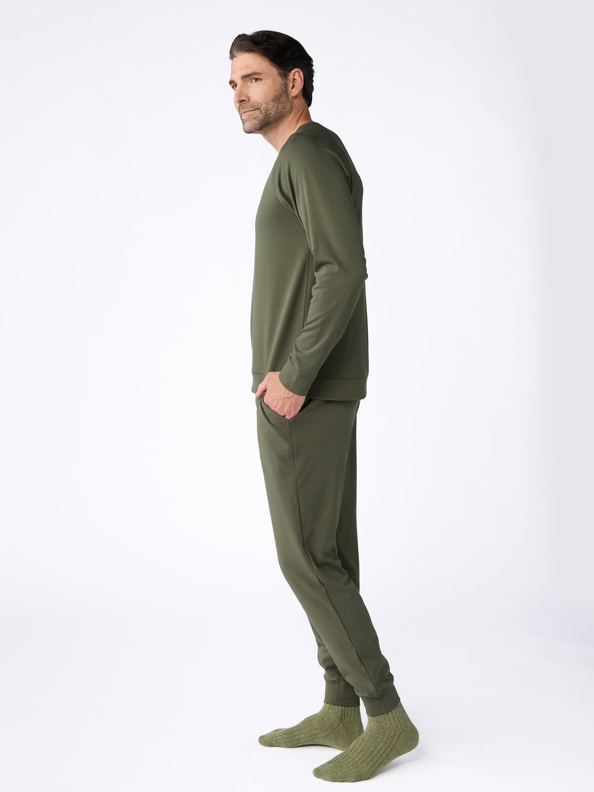 A man with dark hair and a beard stands in profile against a white background, wearing Cozy Earth's Men's Ultra-Soft Bamboo Pullover Crew in olive green, paired with matching joggers and knit socks, his hands tucked into the pockets of his pants. |Color:Olive