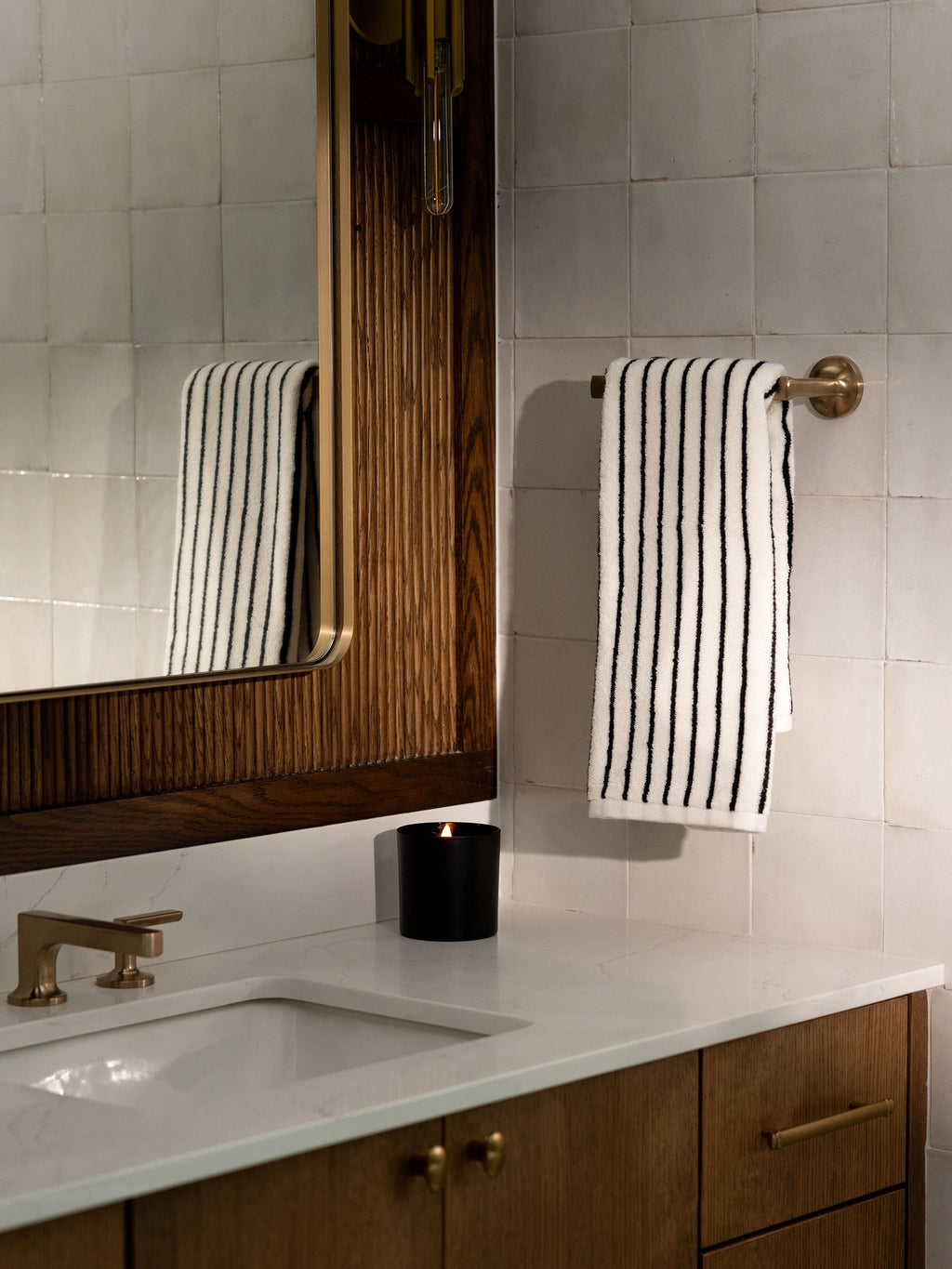 A bathroom scene featuring a white countertop with a sink, brass faucet, and wooden cabinets. A black candle and Luxe Hand Towels by Cozy Earth, hanging on a brass towel bar, are also present. The background consists of white tiled walls and a large wooden-framed mirror. |Color:Black Stripe