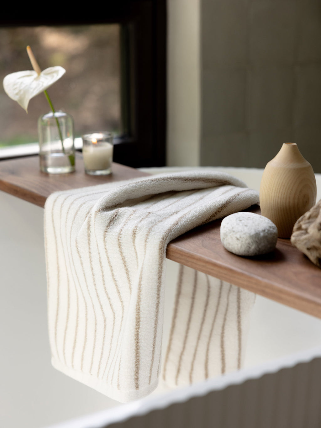 A Luxe Hand Towel by Cozy Earth, featuring a white and beige stripe pattern, hangs over the wooden ledge of a bathtub. Beside the towel on the ledge are a potted flower in a glass vase, a lit candle, a smooth stone, and a wooden decor piece. A window in the background offers a glimpse of greenery outside. |Color:Sand Stripe