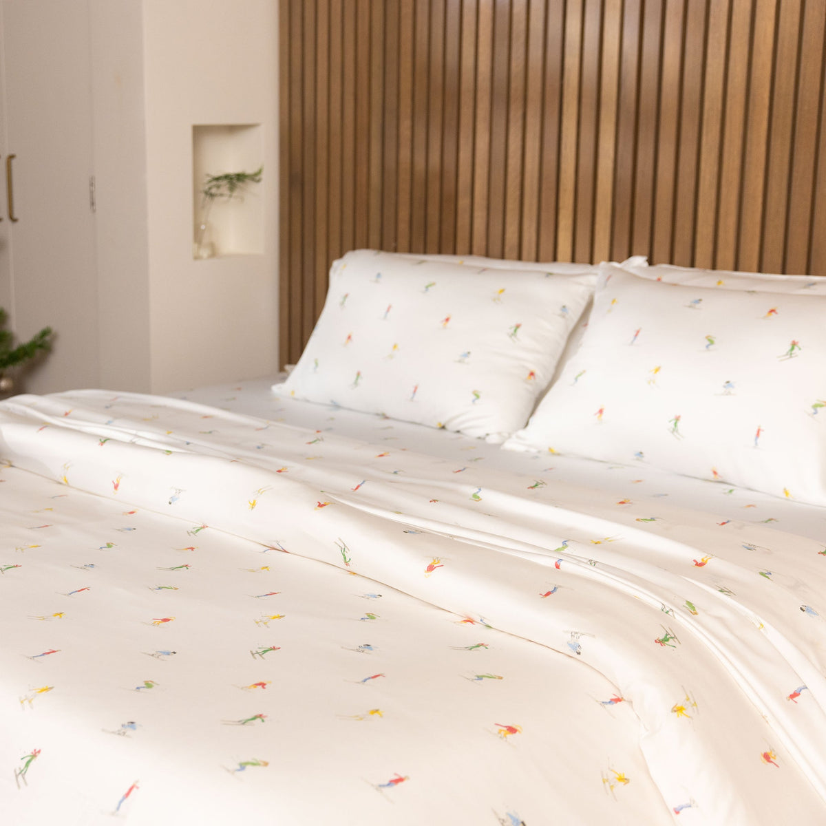 The bed is adorned with a Bamboo Duvet Cover from Cozy Earth, featuring white sheets and pillowcases with a subtle pattern of small, colorful figures. The headboard is crafted from vertical wooden slats, while soft lighting adds to the cozy ambiance. |Color:Skiers