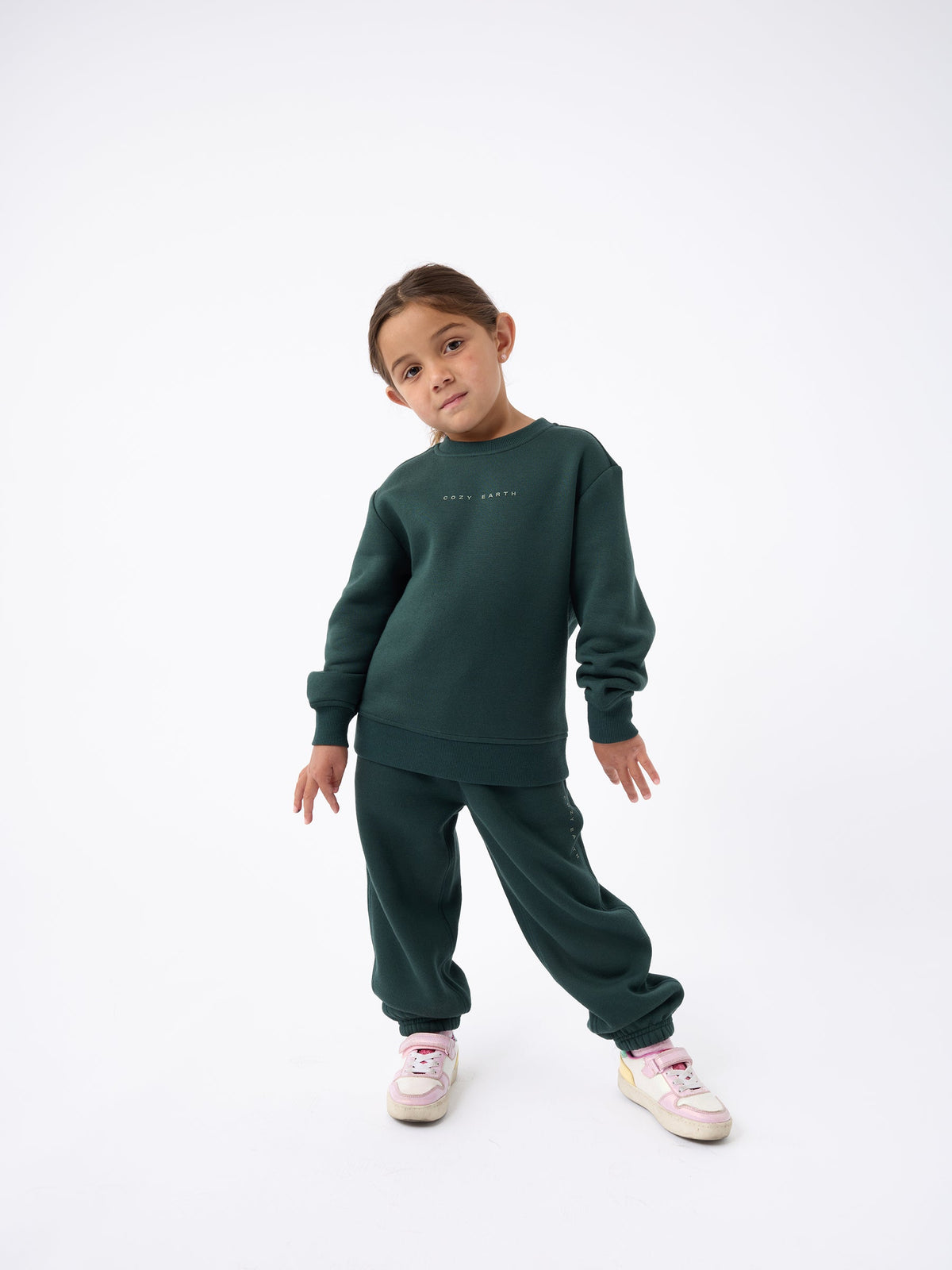 A young child poses against a white background, confidently wearing the Kid's CityScape Crewneck by Cozy Earth in dark green, paired with matching sweatpants and pink and white sneakers, standing with hands slightly bent at the sides. |Color:Spruce