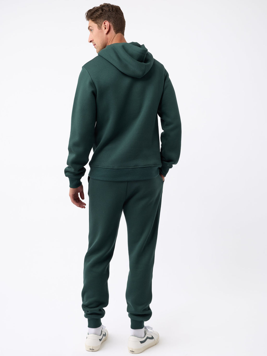 A man wearing the Men's CityScape Jogger by Cozy Earth stands facing away, gazing slightly to the side. He complements his look with white sneakers featuring black accents against a plain white background. |Color:Spruce