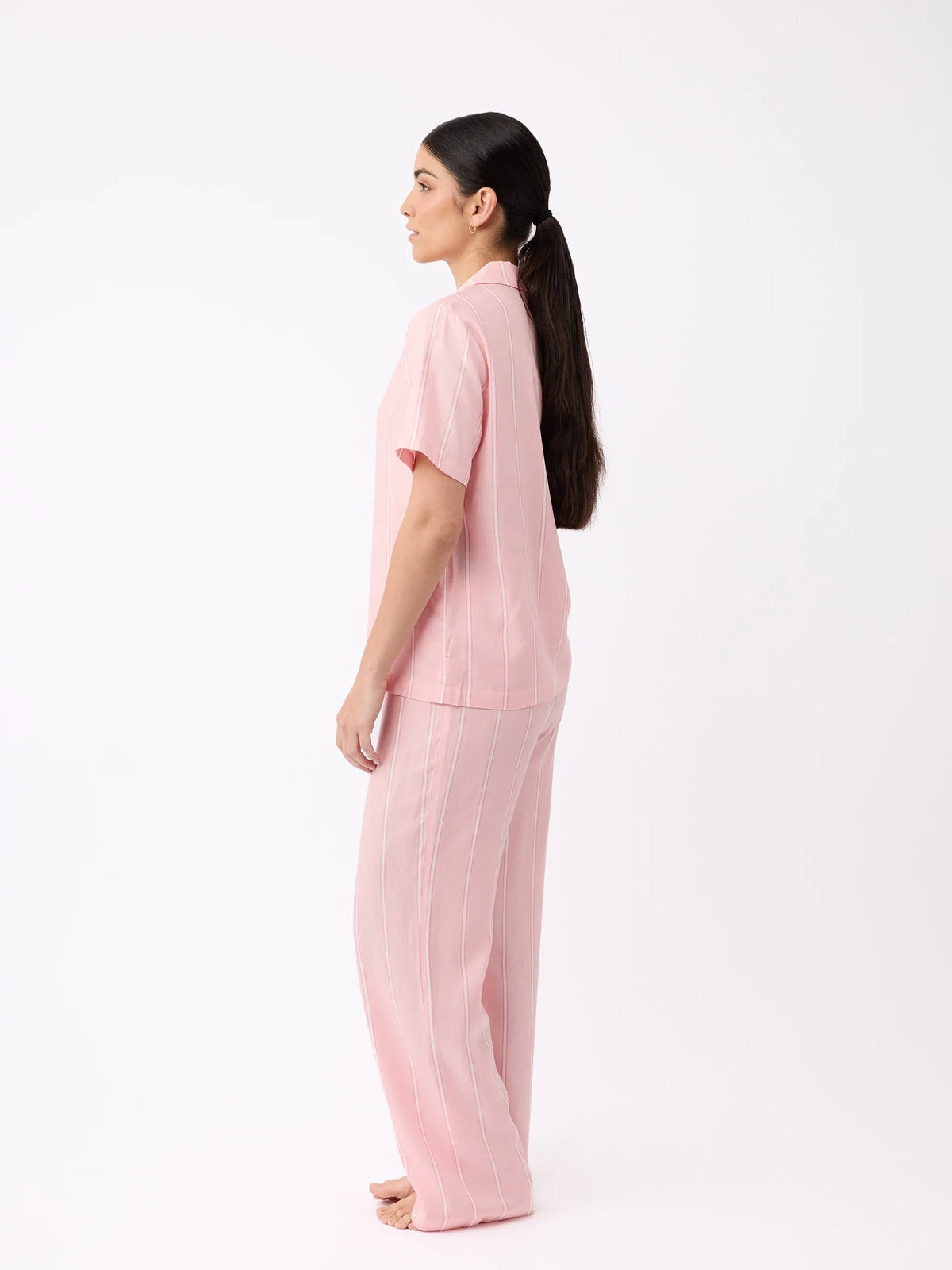A woman with long, dark hair tied in a ponytail stands barefoot and sideways in a white space, wearing Cozy Earth's pink short-sleeved shirt and Women's Soft Woven Pajama Pant. |Color:Sweetheart Stripe