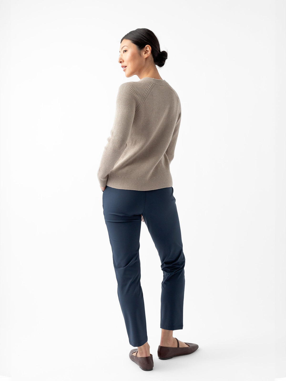 A person is standing against a white background, facing slightly to the side. They are wearing Cozy Earth's Women's Classic Crewneck in beige, dark blue pants, and brown shoes. Their hands are tucked into their pants' pockets, and their dark hair is pulled back in a low bun. |Color:Taupe