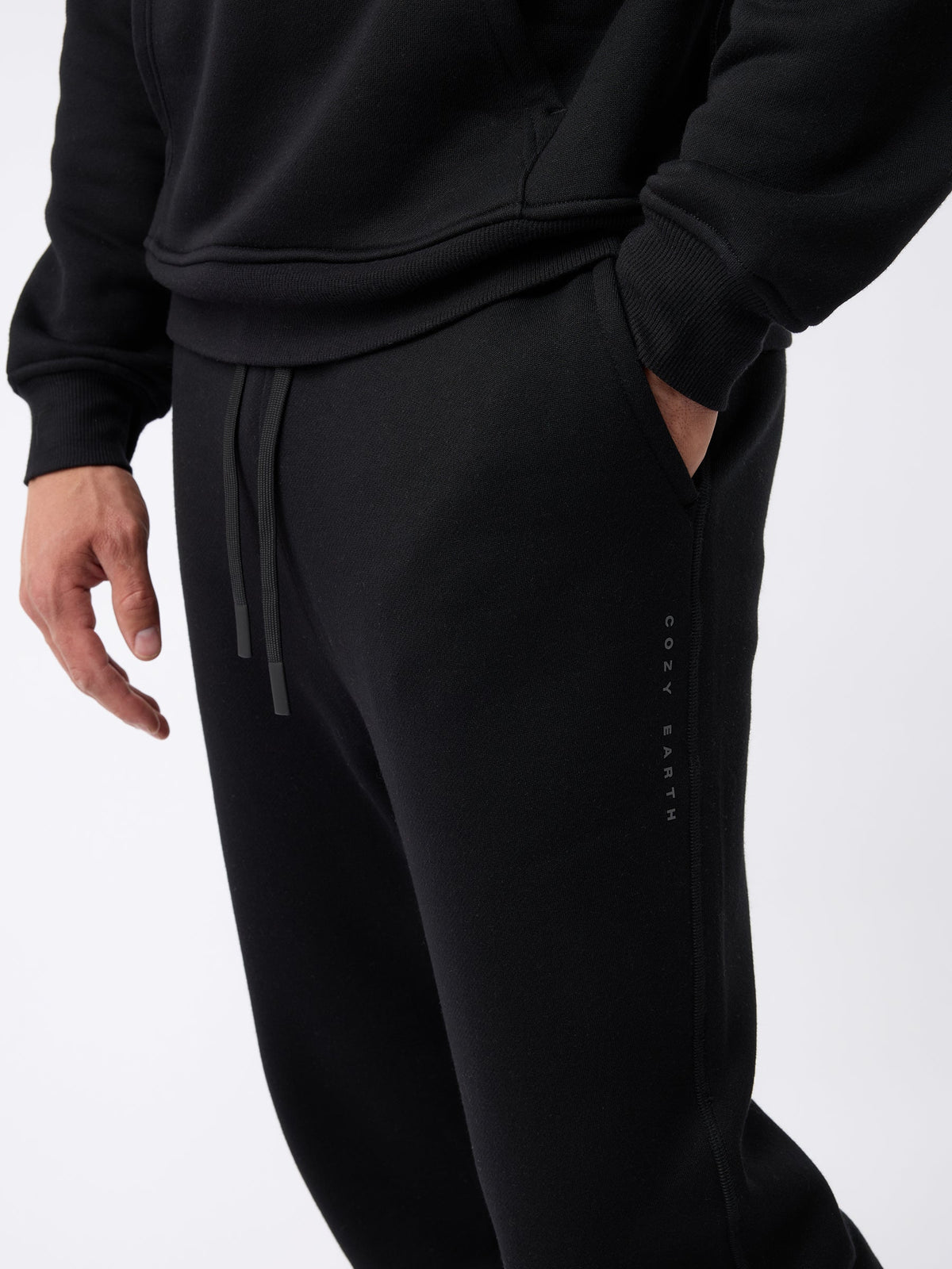 A person is wearing a black hoodie and the Men's CityScape Jogger in black, with their left hand in the jogger's pocket. The brand name "Cozy Earth" is visible on the side of the joggers against a plain white background. |Color:Black