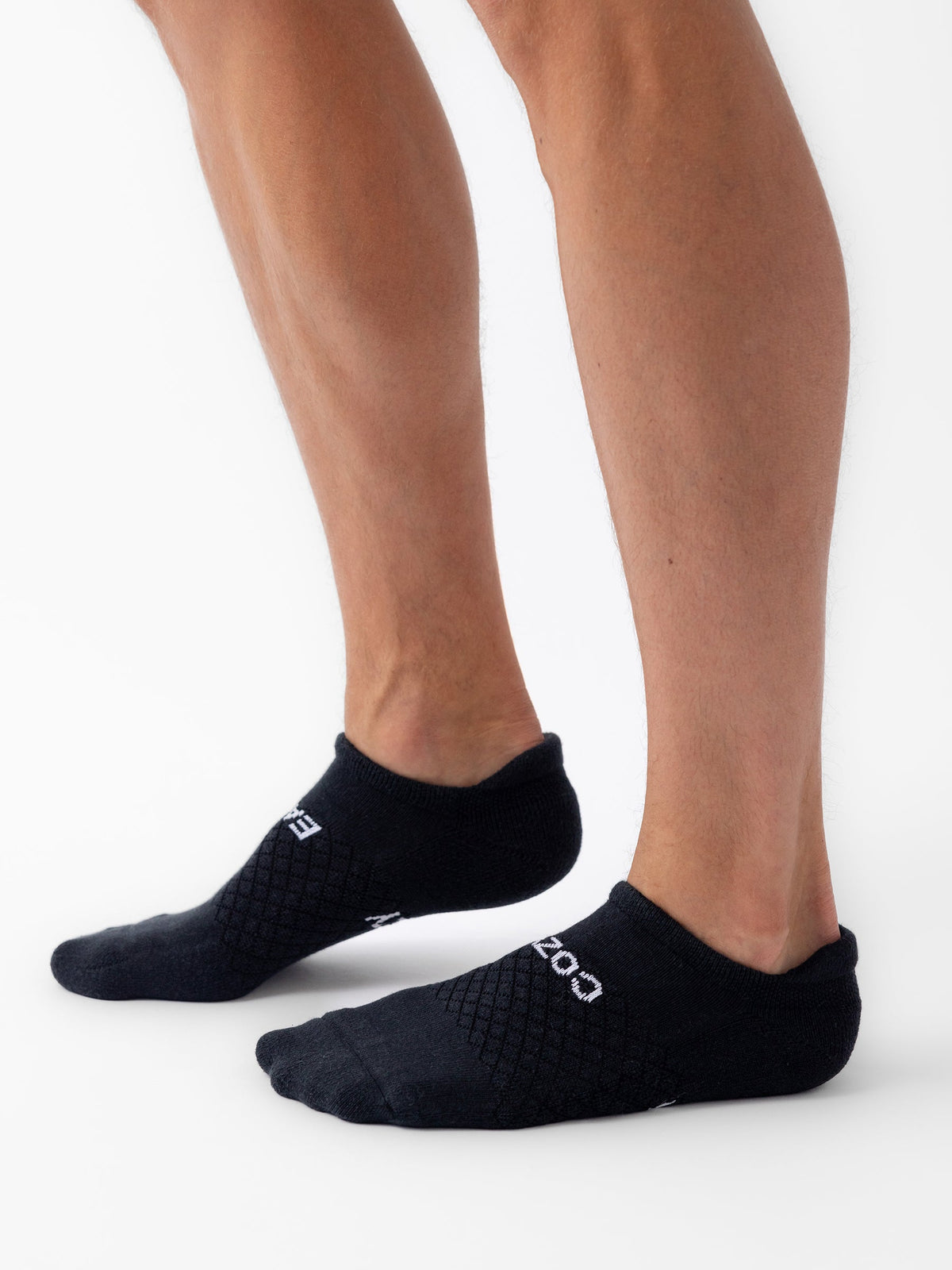 A person wearing black socks from the Essential Ankle Sock 2-Pack by Cozy Earth stands on a white background. The socks feature a textured pattern and the word "COZY" on the sides. The person’s lower legs are visible, and they are standing with one foot slightly in front of the other. |Color:Black|Count:2-Pack