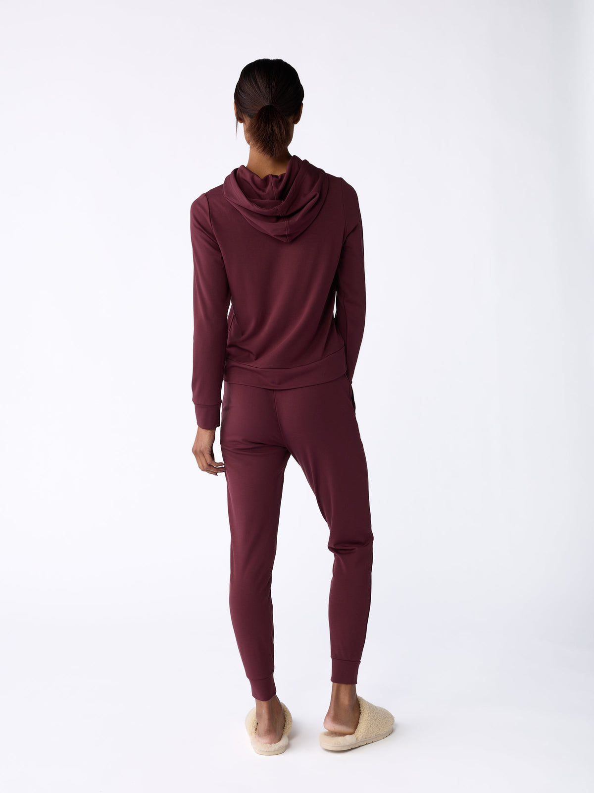 A person stands facing away, wearing Cozy Earth's Women's Bamboo Jogger Pant in maroon and beige slippers. The plain white background accentuates the outfit. |Color:Burgundy