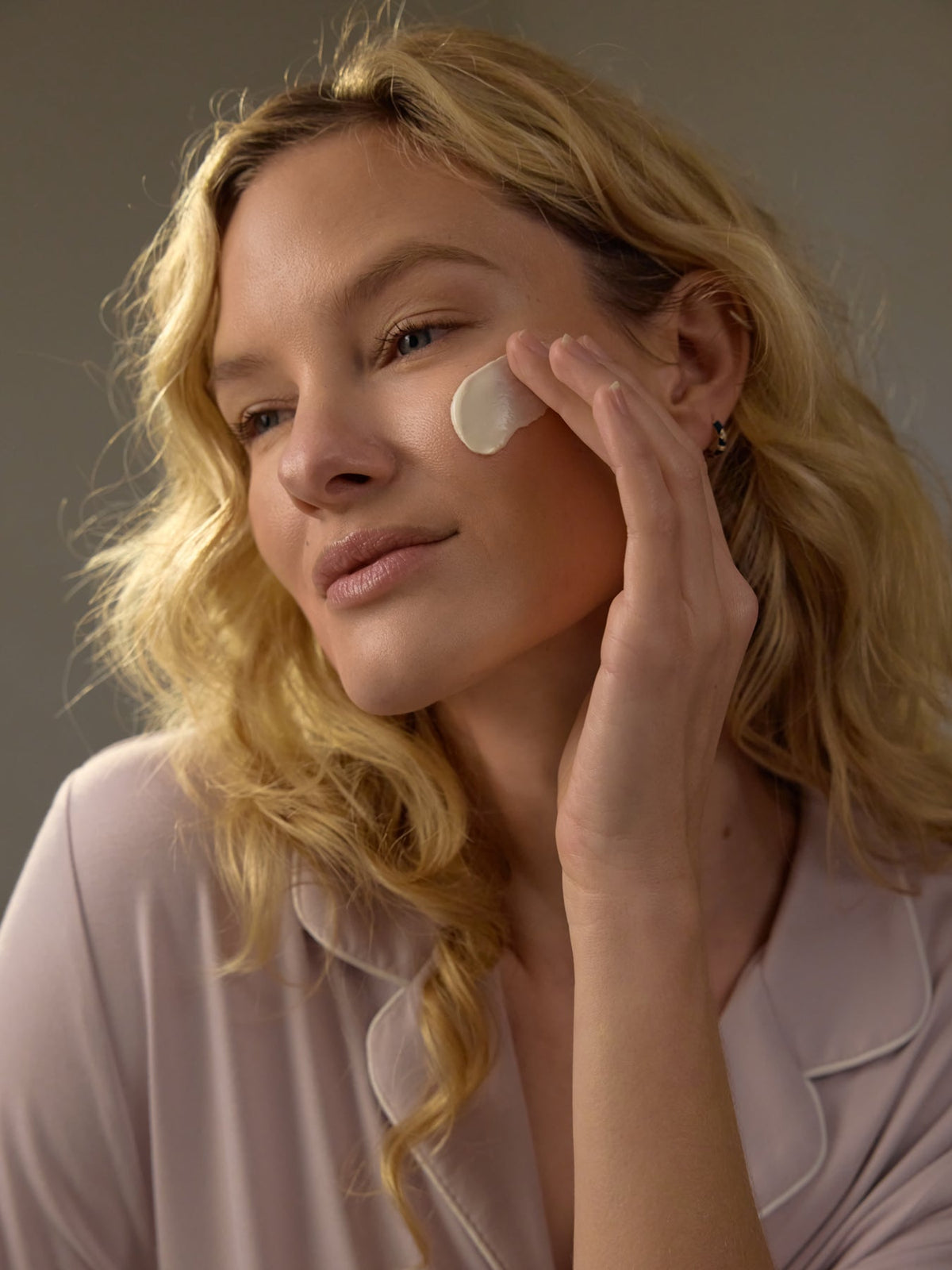 A woman with long blonde hair applies Cozy Earth's Clay Mask to her cheek with her fingers. She wears a light-colored top against a neutral background, appearing content and relaxed. |Size:Mini