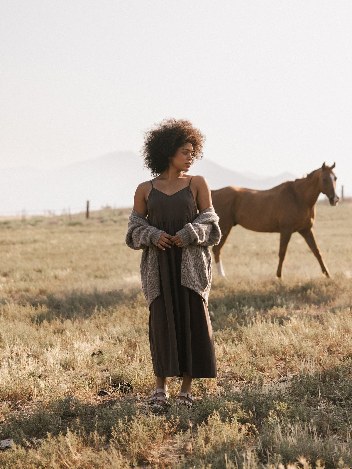 A person with curly hair stands in a grassy field wearing Cozy Earth's Women's Sunset Gathered Dress and a light gray cardigan, with eyes gazing downward. In the background, a brown horse walks casually. Misty mountains are visible in the distance under a pale sky. |Color:Cacao
