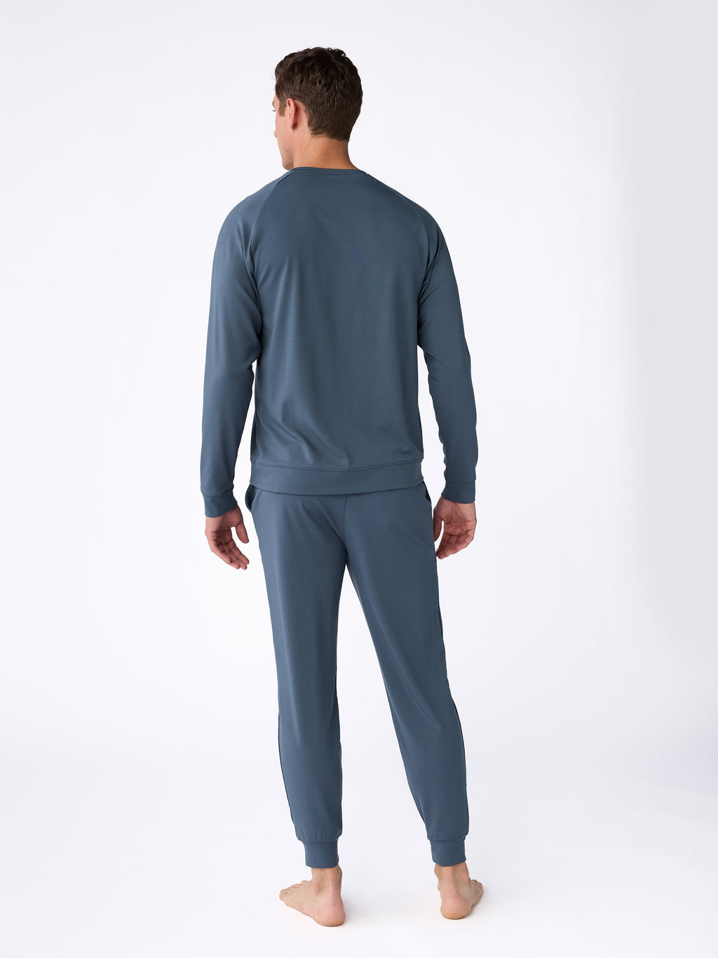 A person wearing a blue Men's Ultra-Soft Bamboo Pullover Crew by Cozy Earth stands barefoot on a white background, facing away from the camera. |Color:Dawn