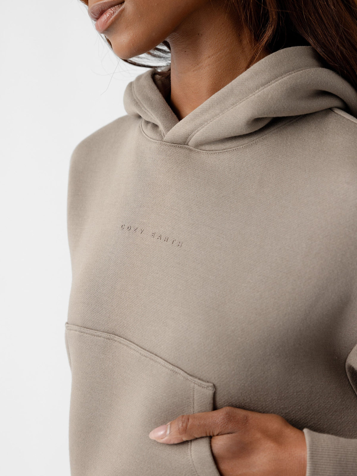 A close-up shot captures a person wearing the Women's CityScape Cropped Hoodie by Cozy Earth. The lower part of their face, neck, and upper torso are shown. This beige hoodie features a front pocket and text embroidered on the chest. The individual’s hand rests inside the pocket, against a plain white background. |Color:Ebony