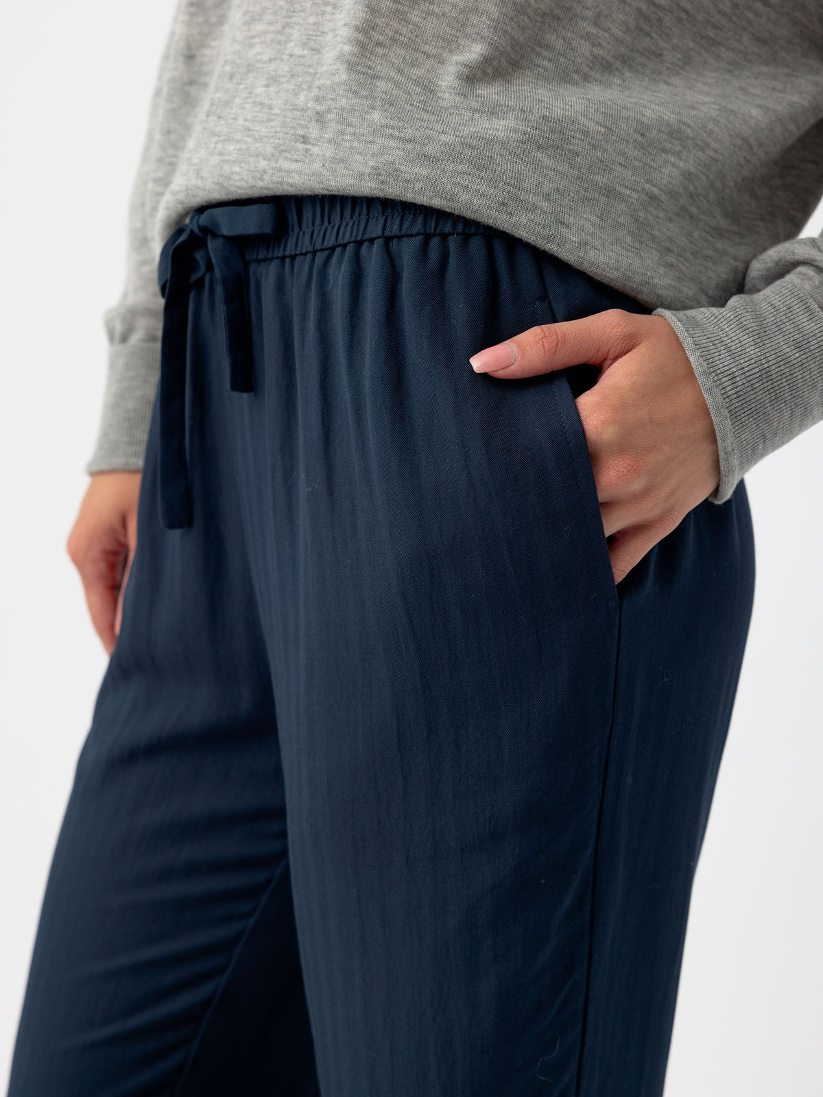 A person wearing a grey sweater and Cozy Earth's Women's Sunset Cropped Pant in dark blue, featuring a loose fit and an elastic waistband, stands with one hand in their pocket. The image highlights the midsection and part of the arm, showcasing the relaxed fit and texture of the clothing. |Color:Eclipse
