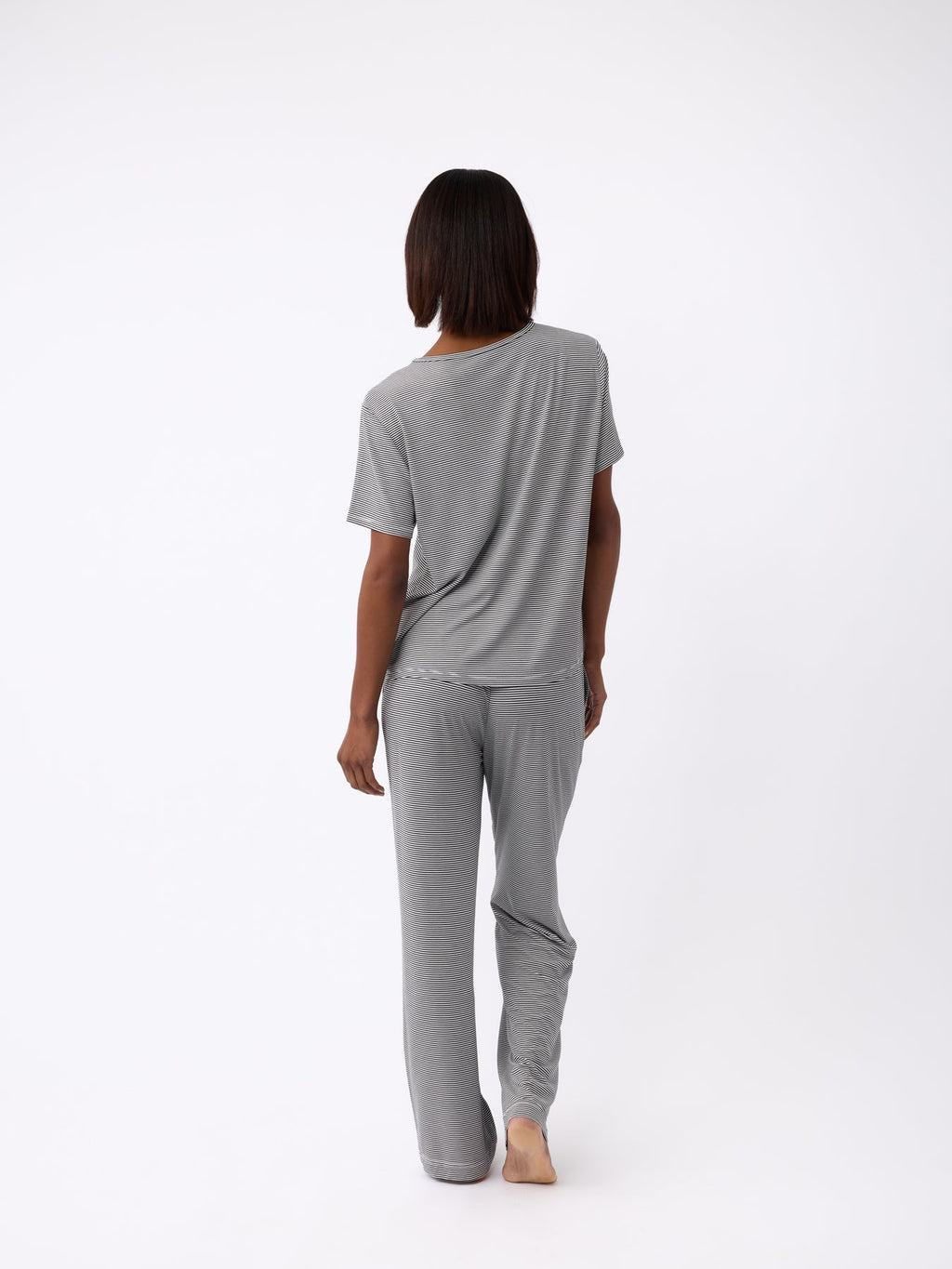 A person with shoulder-length hair stands barefoot, facing away, wearing a loose-fitting gray t-shirt along with Cozy Earth's Women's Bamboo Stretch-Knit Pant. The background is plain white. |Color:Eclipse Mini Stripe