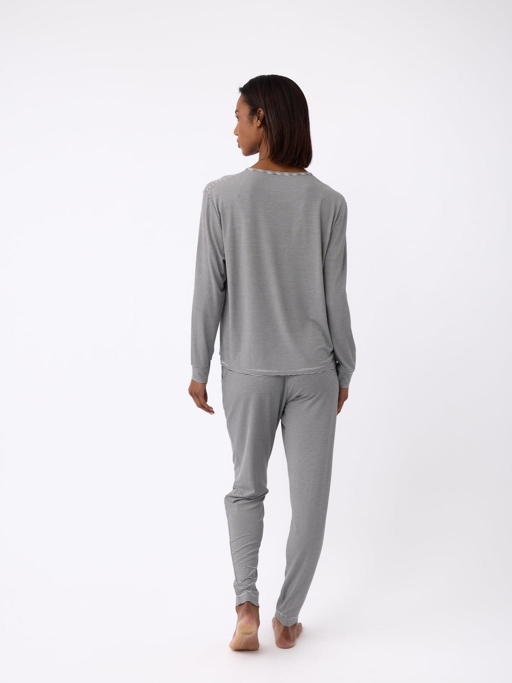 A person stands barefoot, facing away, wearing Cozy Earth's Women's Bamboo Stretch-Knit Tapered Pant in gray loungewear on a white background. The outfit includes a long-sleeve top and pants. |Color:Eclipse Mini Stripe