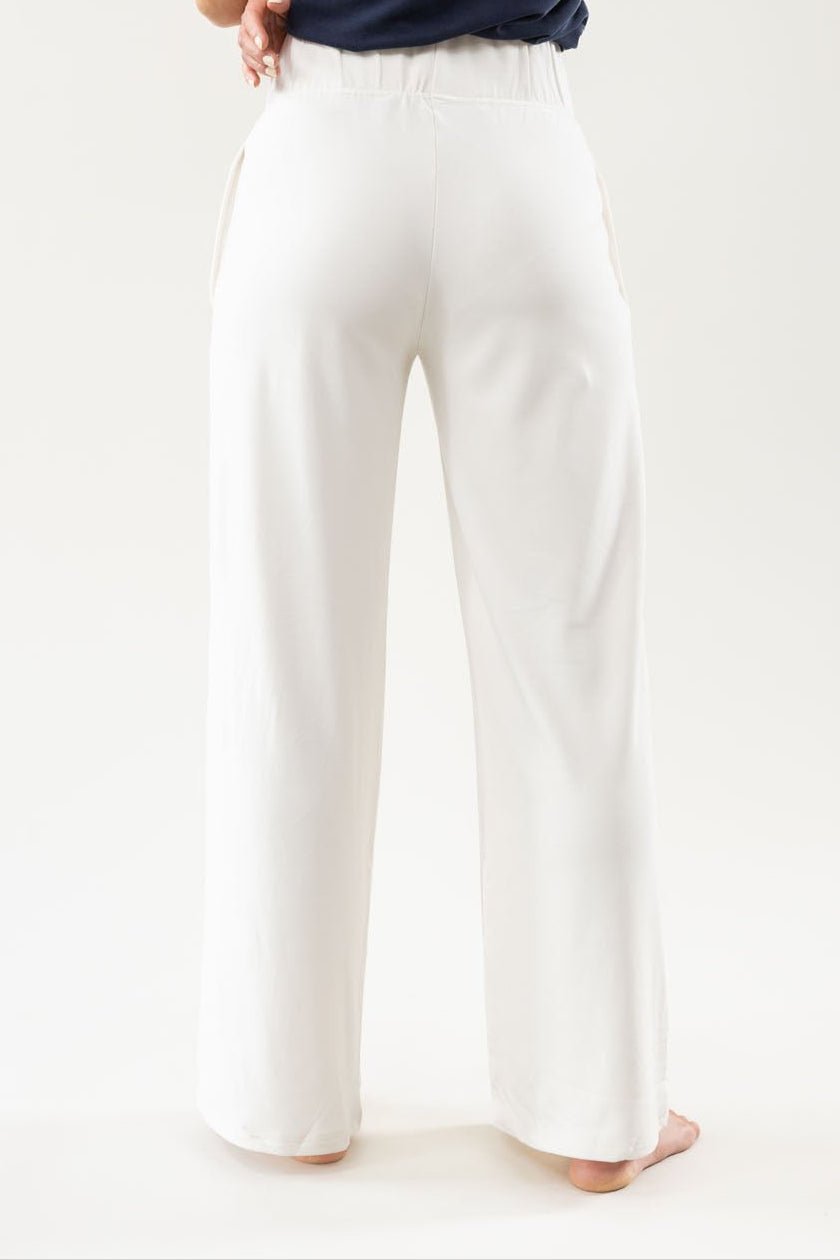 A person stands barefoot with their back to the camera, wearing a navy top and loose-fitting white pants from the Women's Ultra-Soft Bamboo Wide Leg Pull On Pant & Pullover Set by Cozy Earth. The background is plain and light-colored. |Color:Ivory