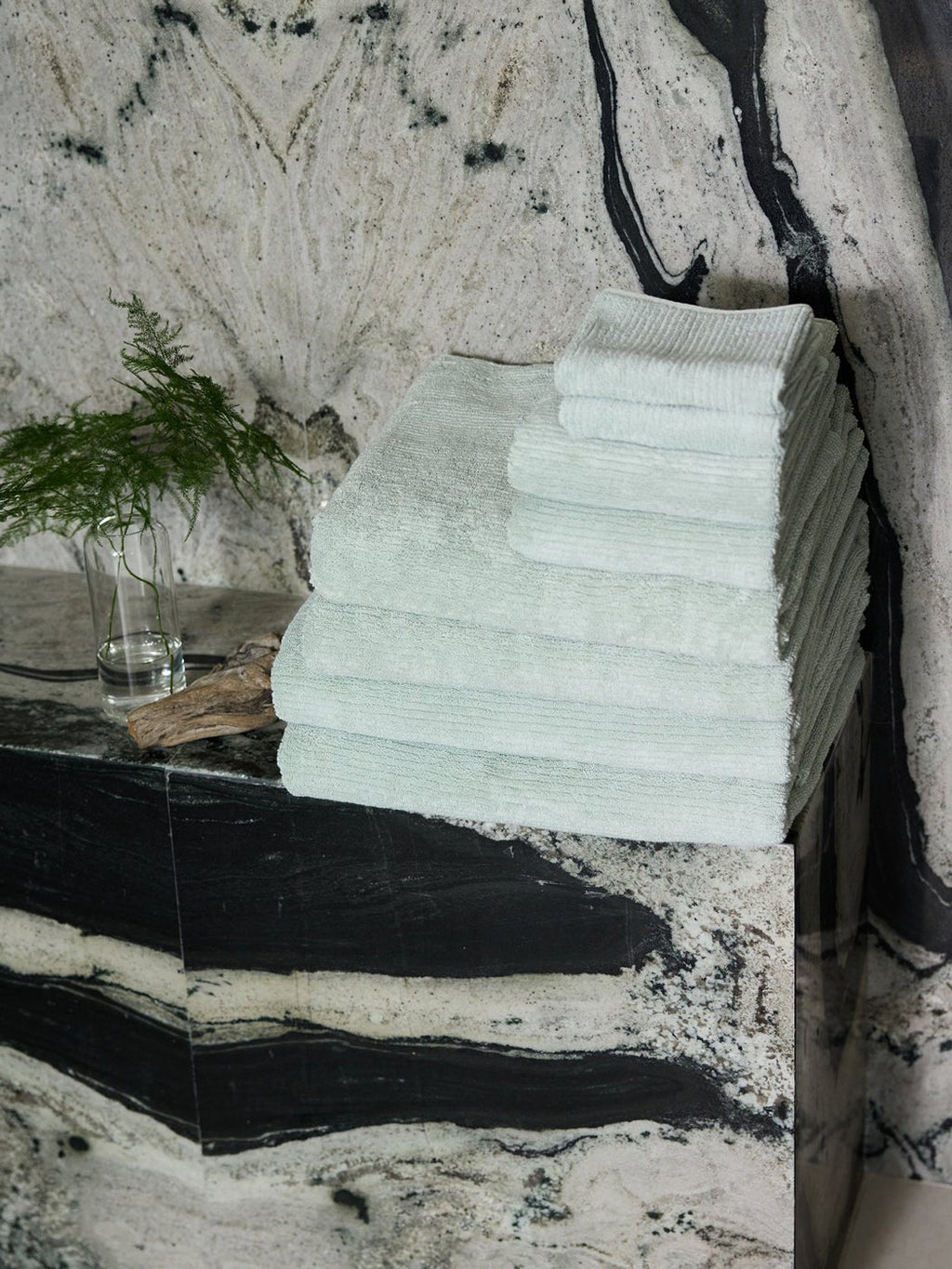 The Complete Ribbed Terry Bath Bundle by Cozy Earth is stacked neatly on a black and white marble counter beside a vase with a green fern, set against a matching marble wall.|Color:Laurel