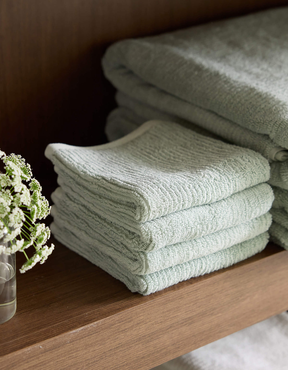 A neatly folded stack of Cozy Earth's Ribbed Terry Washcloths in pale green is placed on a wooden shelf. Beside the washcloths, a small clear glass with a few delicate white flowers in water adds a touch of elegance to the display. |Color:Laurel