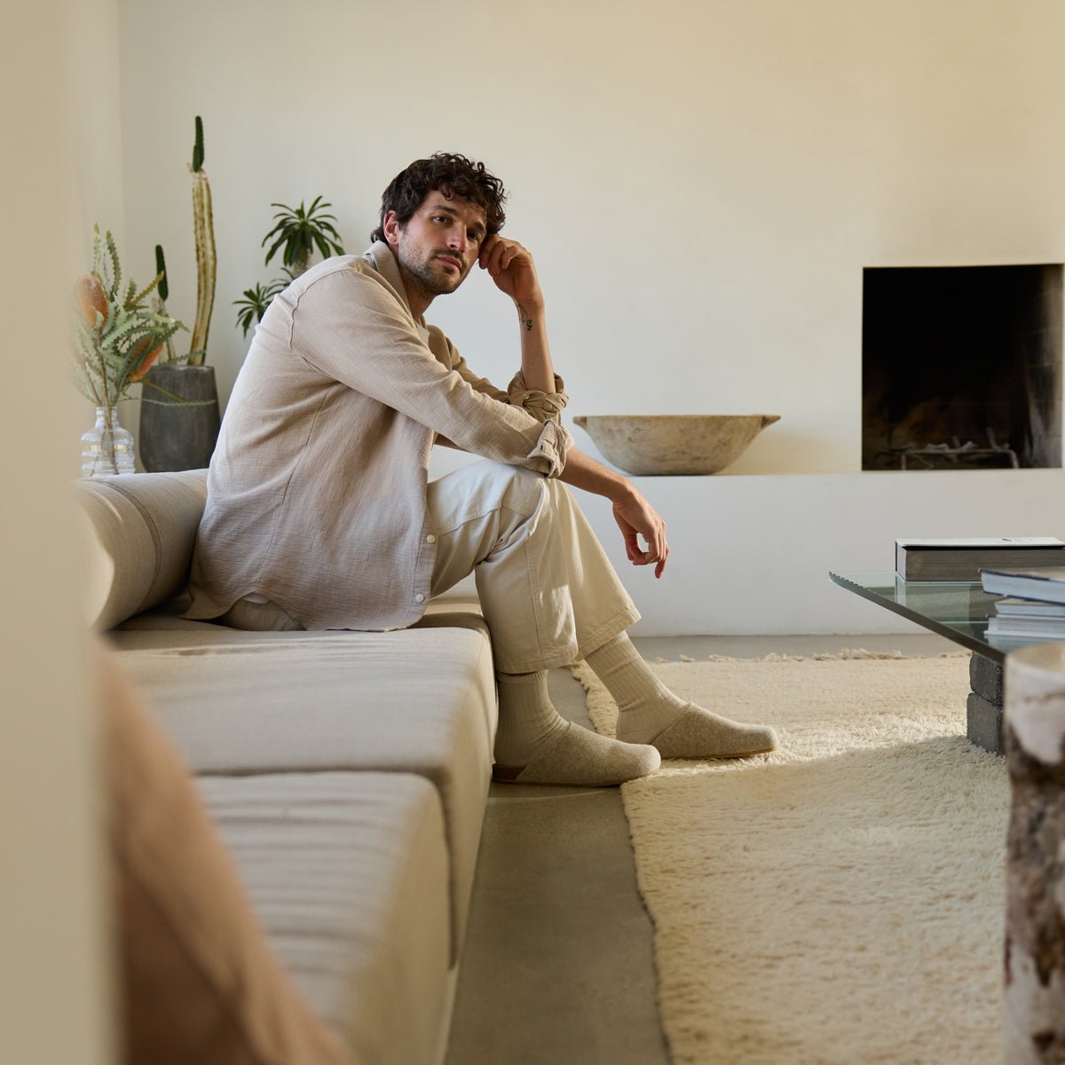 A man with curly hair in a light beige shirt, cream pants, and socks sits on a minimalist beige sofa in a neutral-toned living room. A cactus, other plants, a fireplace, and a glass coffee table are in the background. He's wearing Cozy Earth's Lakehouse Clog shoes. |Color:Oat