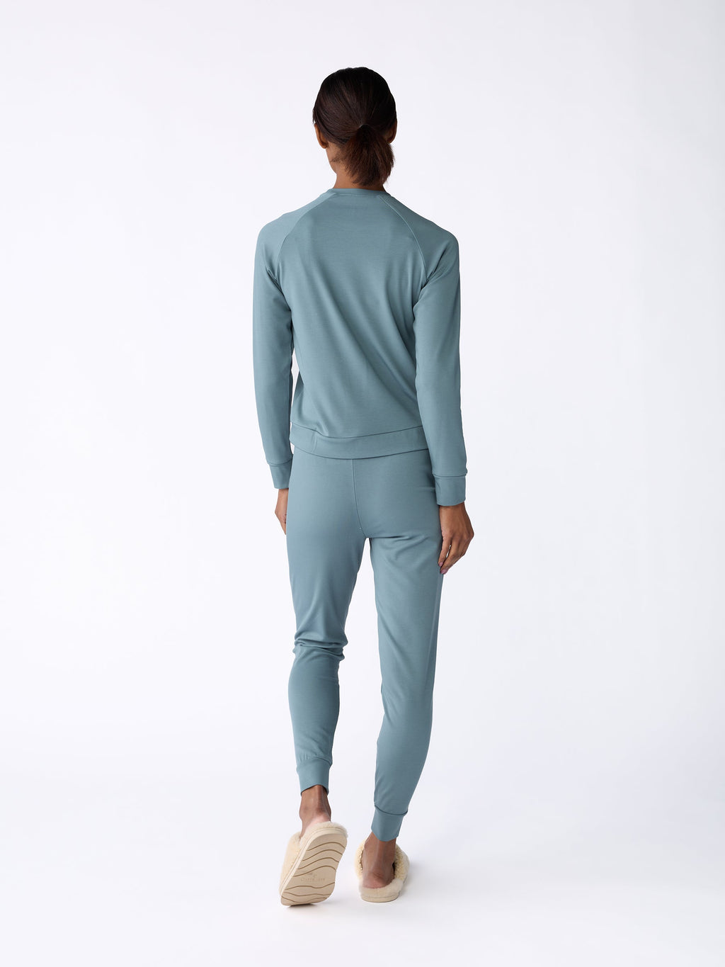A woman is seen facing away, dressed in a matching blue-green tracksuit, featuring the Women's Bamboo Jogger Pant by Cozy Earth, complemented by beige slip-on shoes. The plain white background highlights the clothing ensemble. |Color:Oceanside