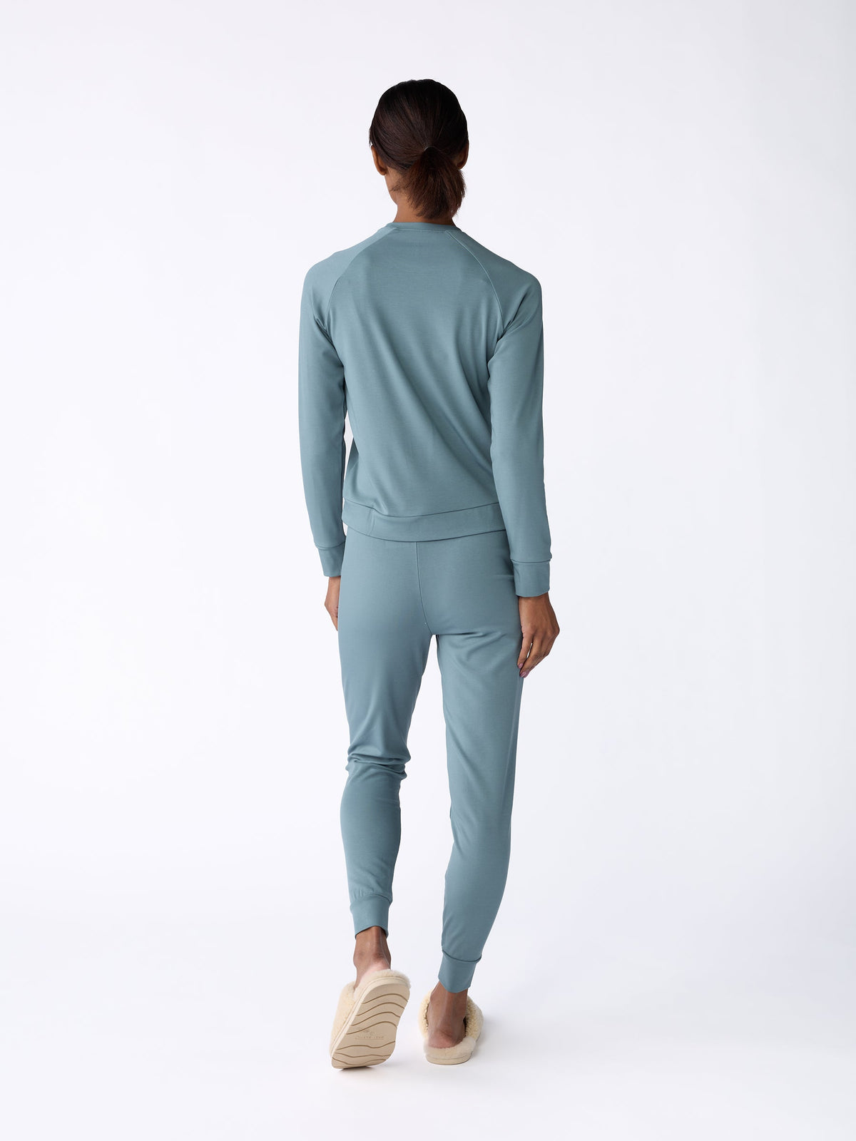 A woman is seen facing away, dressed in a matching blue-green tracksuit, featuring the Women's Bamboo Jogger Pant by Cozy Earth, complemented by beige slip-on shoes. The plain white background highlights the clothing ensemble. |Color:Oceanside